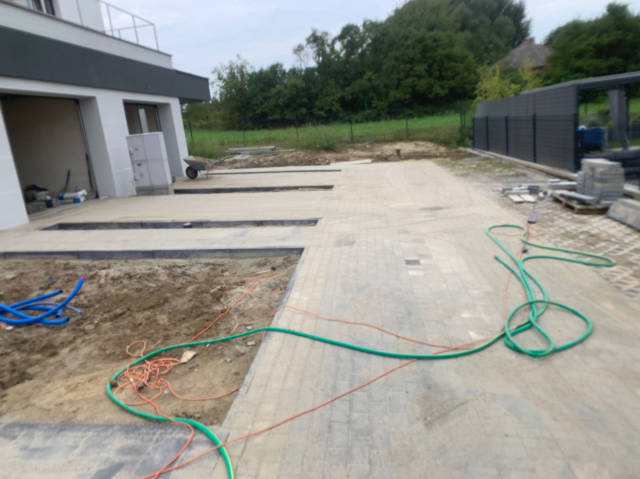 Fragment posesji w trakcie prac brukarskich: częściowo ułożona kostka, przygotowane podłoże z widocznymi wykopami, zwoje węży ogrodowych, paleta z kostką w tle, nowoczesny budynek z garażem w tle.