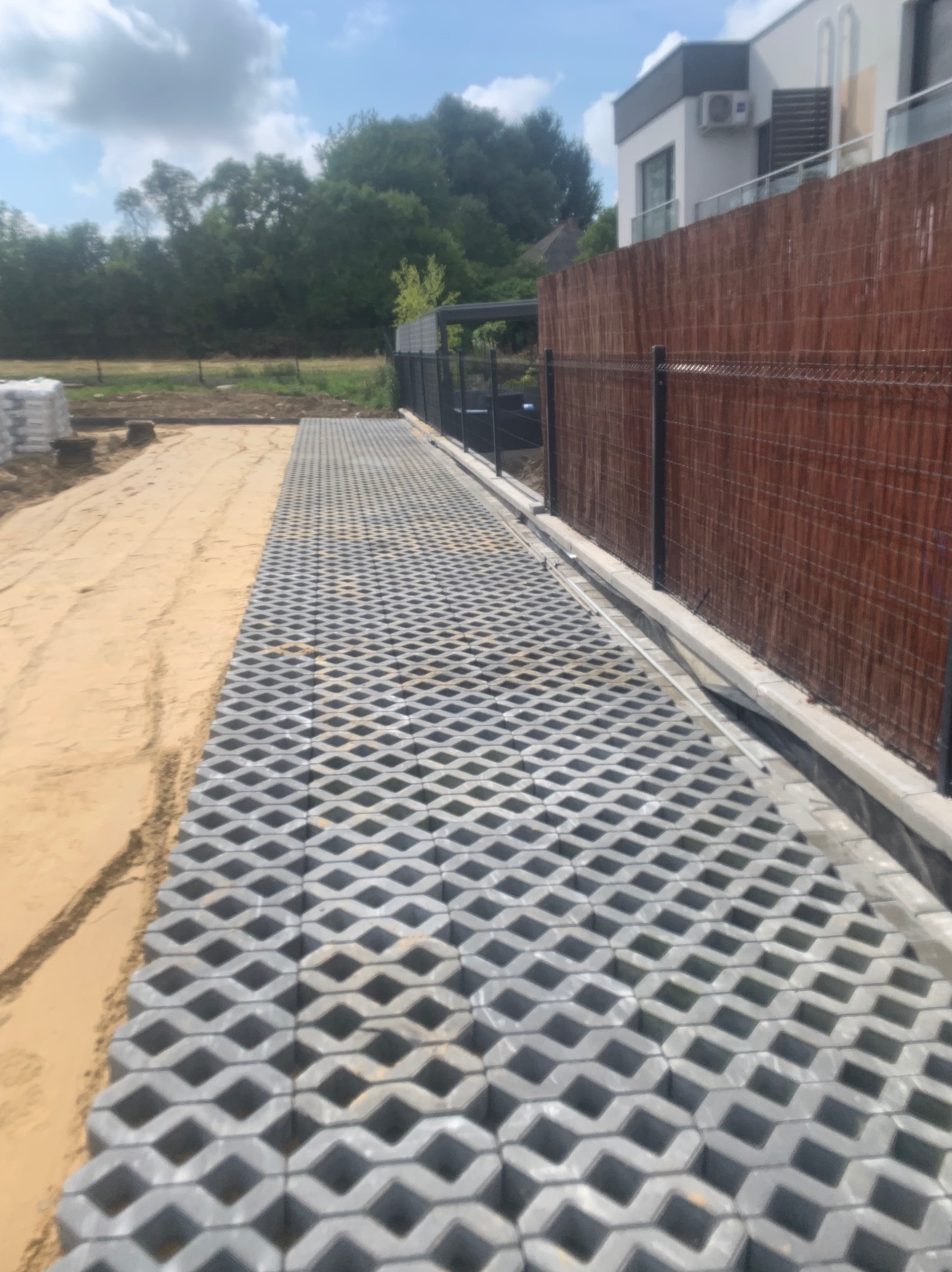 Ułożona szara ażurowa nawierzchnia na piaskowym podłożu w Rzeszowie, widok z góry, obok ogrodzenie z brązowej wikliny i siatki.