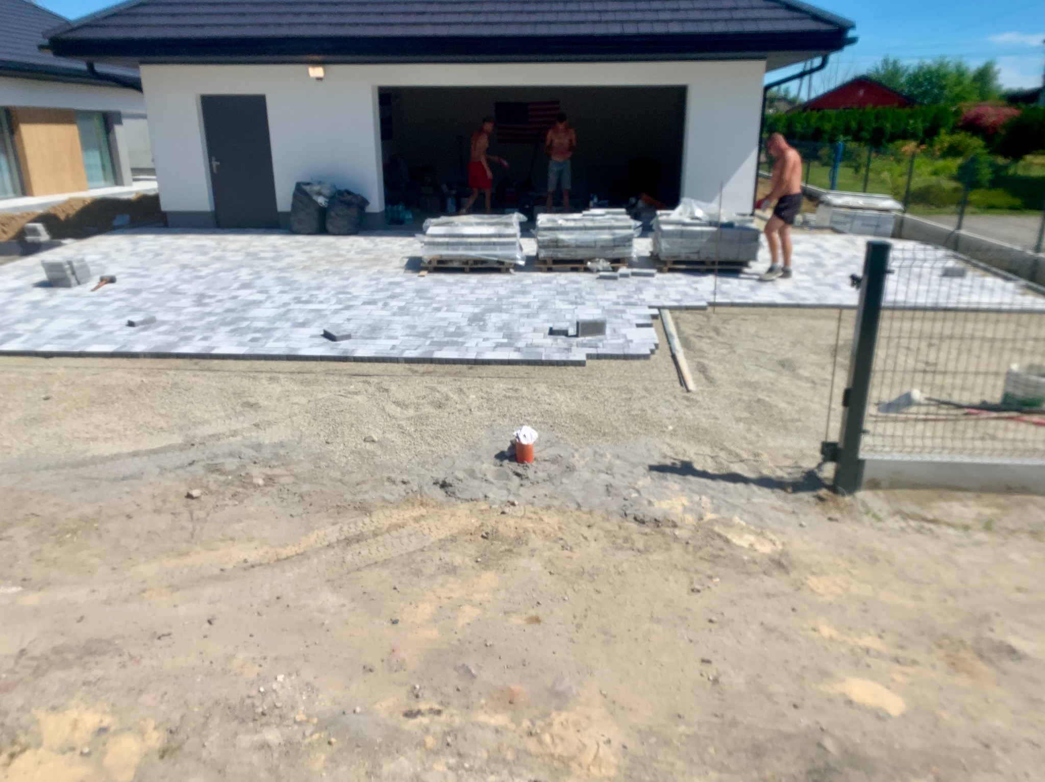 Prace brukarskie w toku: częściowo ułożona szara kostka brukowa przed garażem, palety z kostką, pracownicy bez koszulek w tle, widoczny fragment ogrodzenia i przygotowany teren.