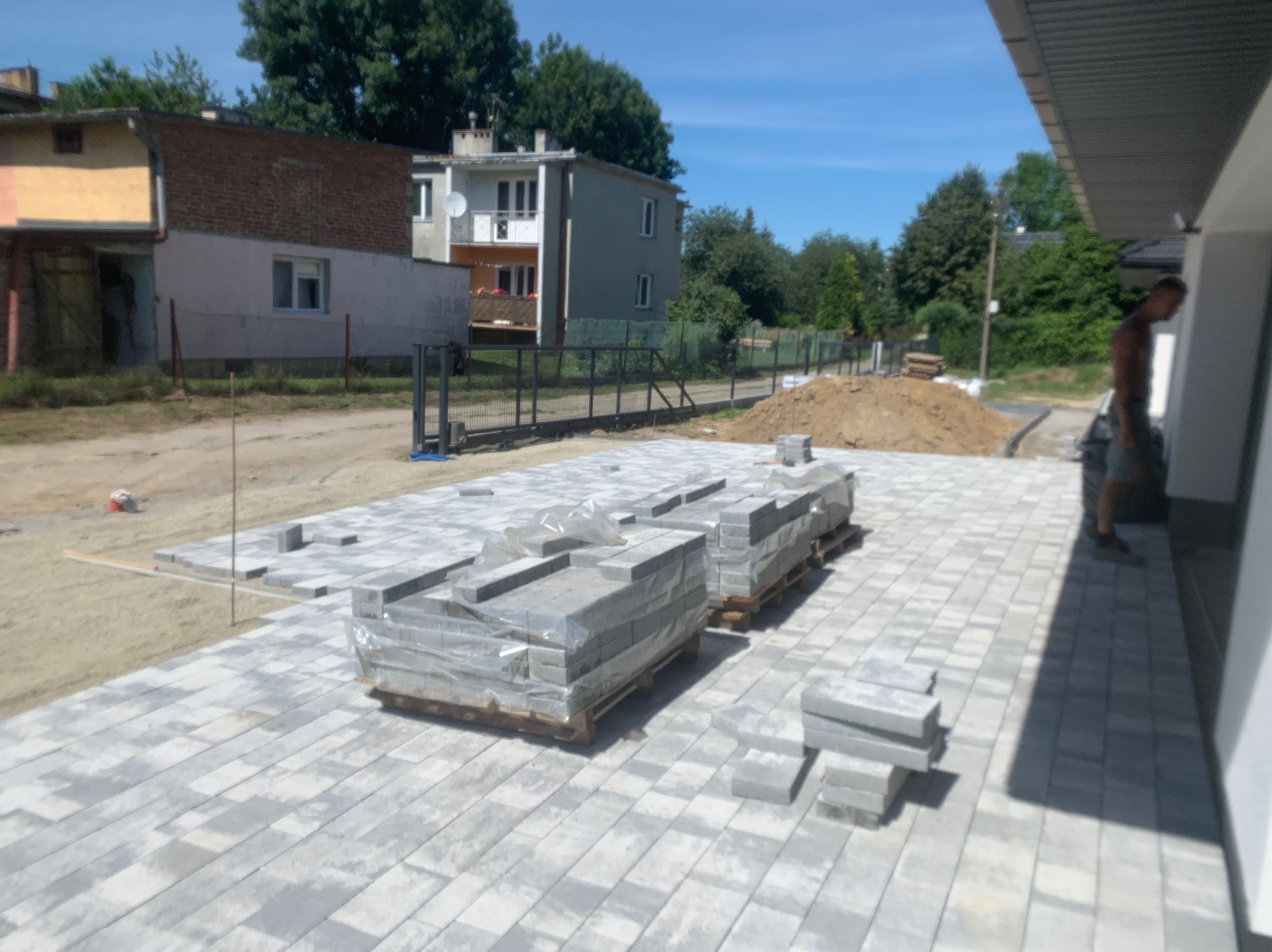 Nierówne ułożenie szarej kostki brukowej na piasku, palety z kostką w folii, przygotowany teren pod dalsze prace, widok na domy w tle.