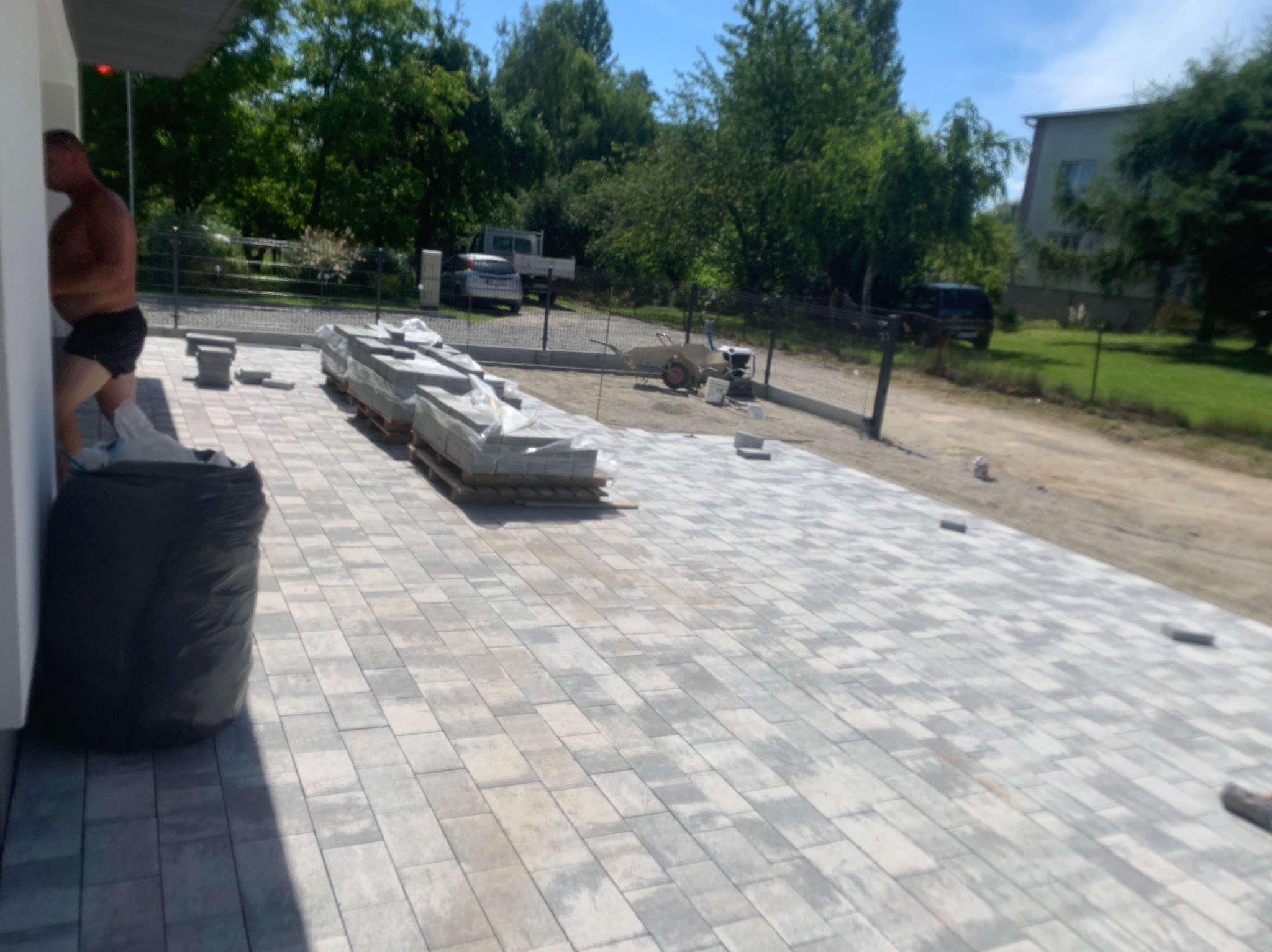 Położona szara kostka brukowa na posesji, paleta z zapakowaną kostką w tle, widoczny pracownik przy ścianie budynku.