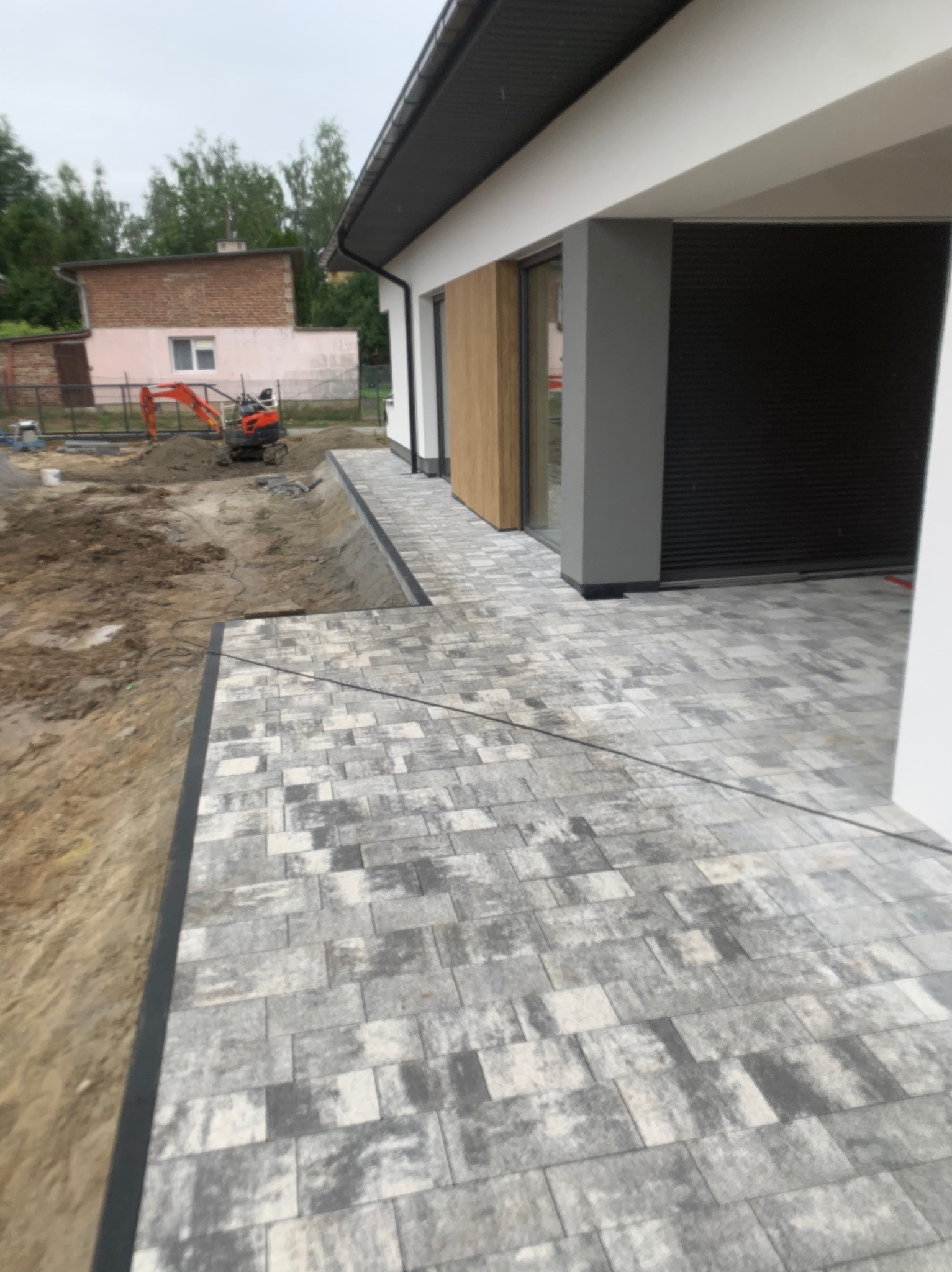 Ułożona kostka brukowa w odcieniach szarości przed nowoczesnym domem, w tle minikoparka przygotowująca teren.