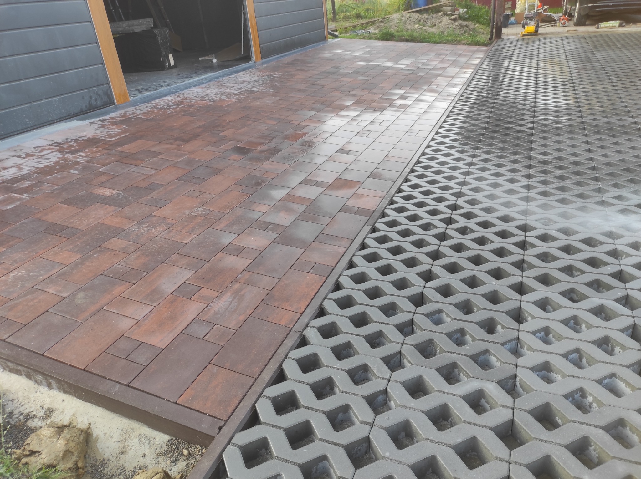 Ułożona, częściowo mokra, brązowo-szara kostka brukowa sąsiadująca z szarymi ażurowymi płytami drogowymi na podjeździe przed garażem.