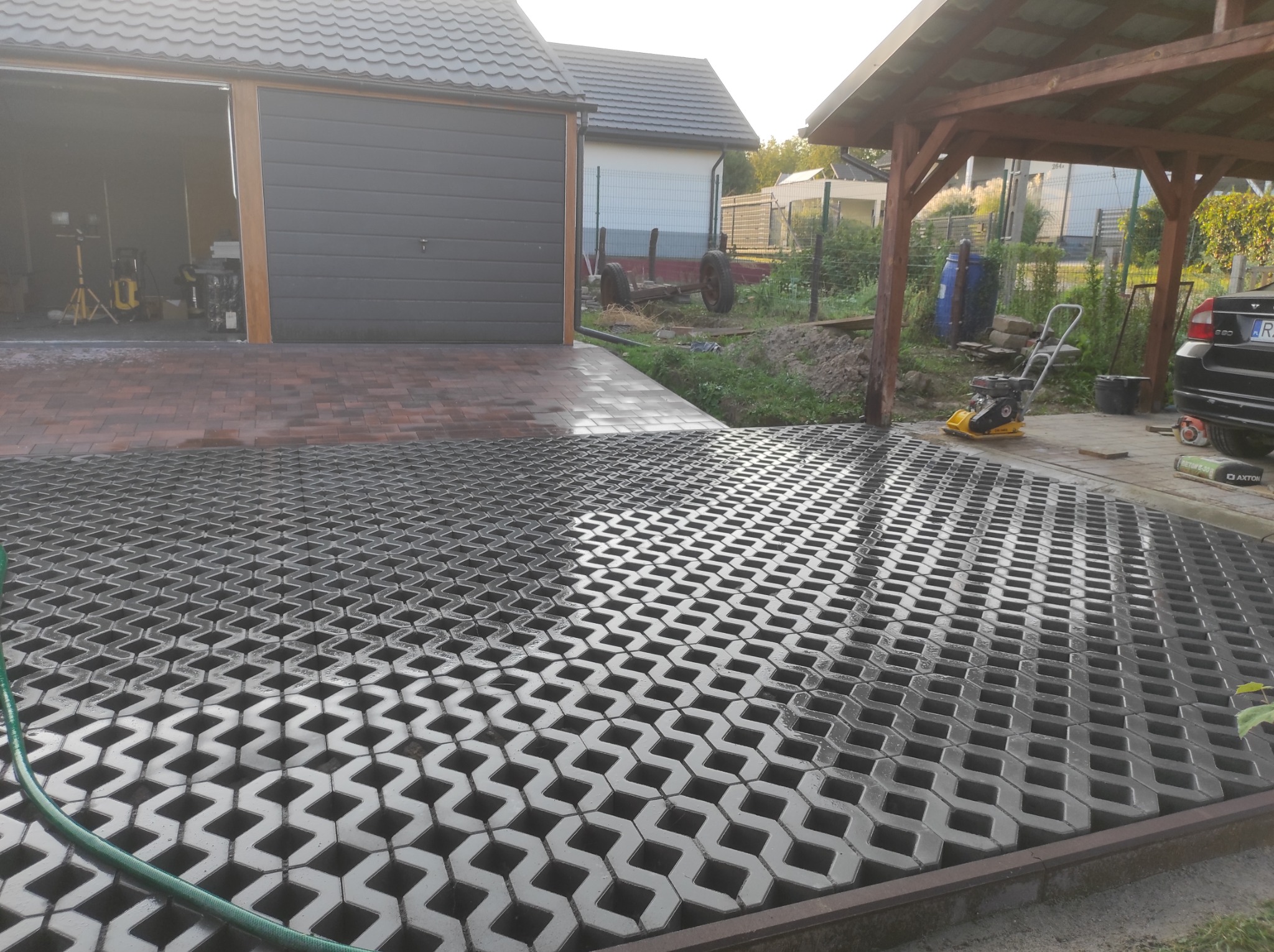 Częściowo ułożona nawierzchnia z ażurowych betonowych płyt, obok garaż z szarą bramą i wiata, w tle dom z szarą dachówką, widoczna zagęszczarka wibracyjna.