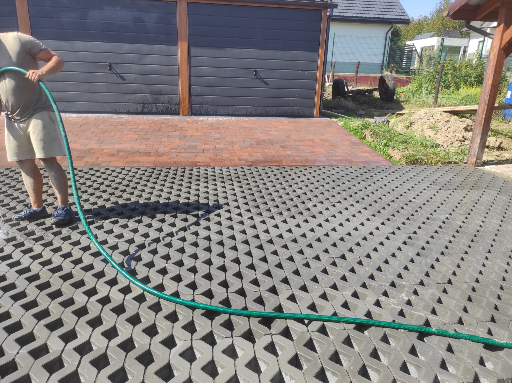 Czyszczenie powierzchni utwardzonej z ażurowych płyt betonowych przy użyciu węża ogrodowego, widoczny fragment garażu z dwoma bramami.