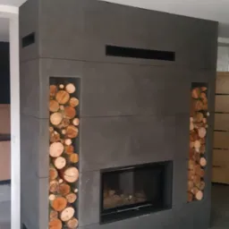 Nowoczesny kominek z obudową z szarego betonu architektonicznego, z wnękami wypełnionymi drewnem opałowym po bokach, umieszczony w salonie z szarą podłogą.