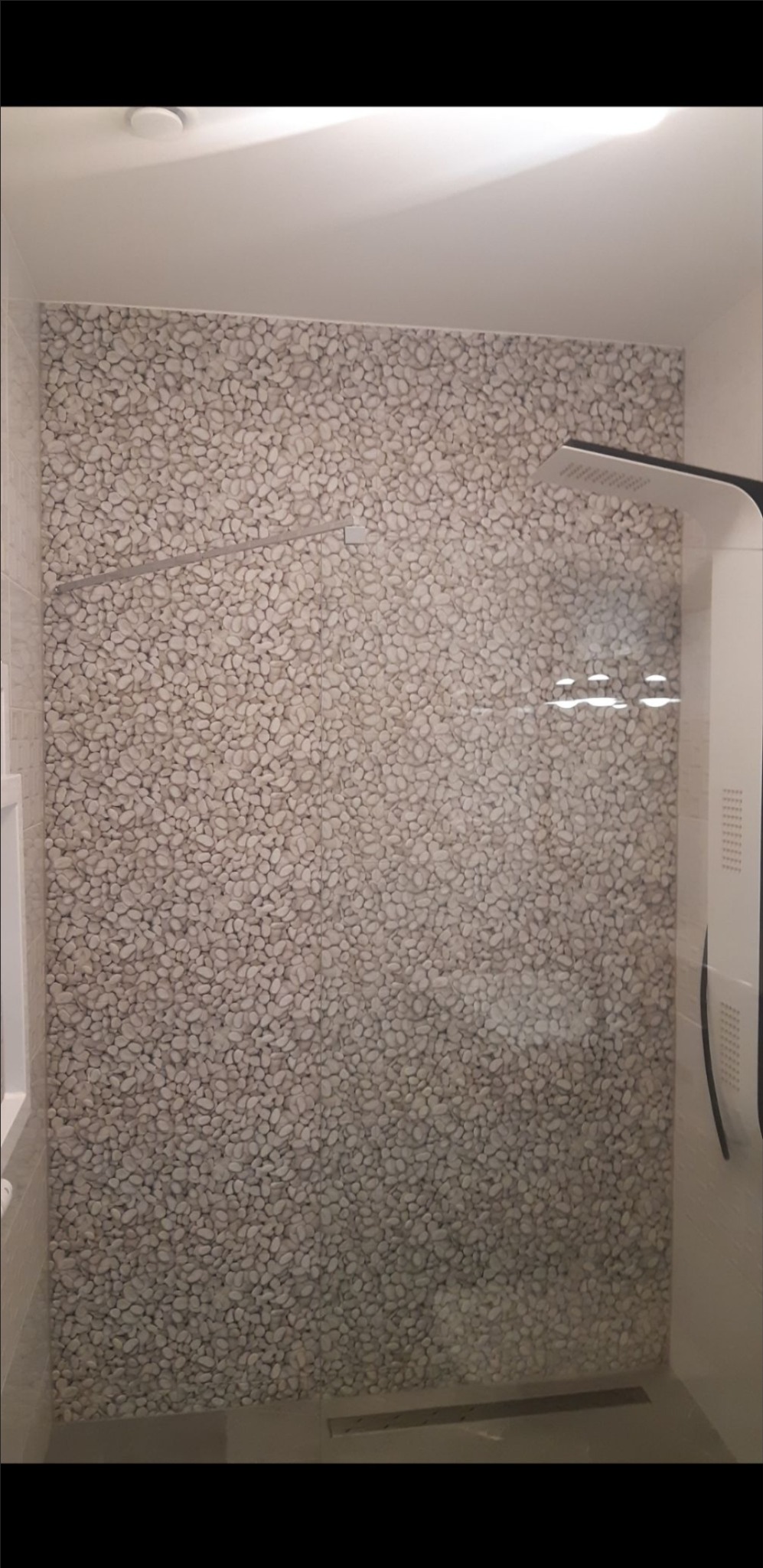 Kabina prysznicowa z wykończeniem ściany imitującym kamyki, nowoczesna słuchawka prysznicowa, odpływ liniowy w podłodze.