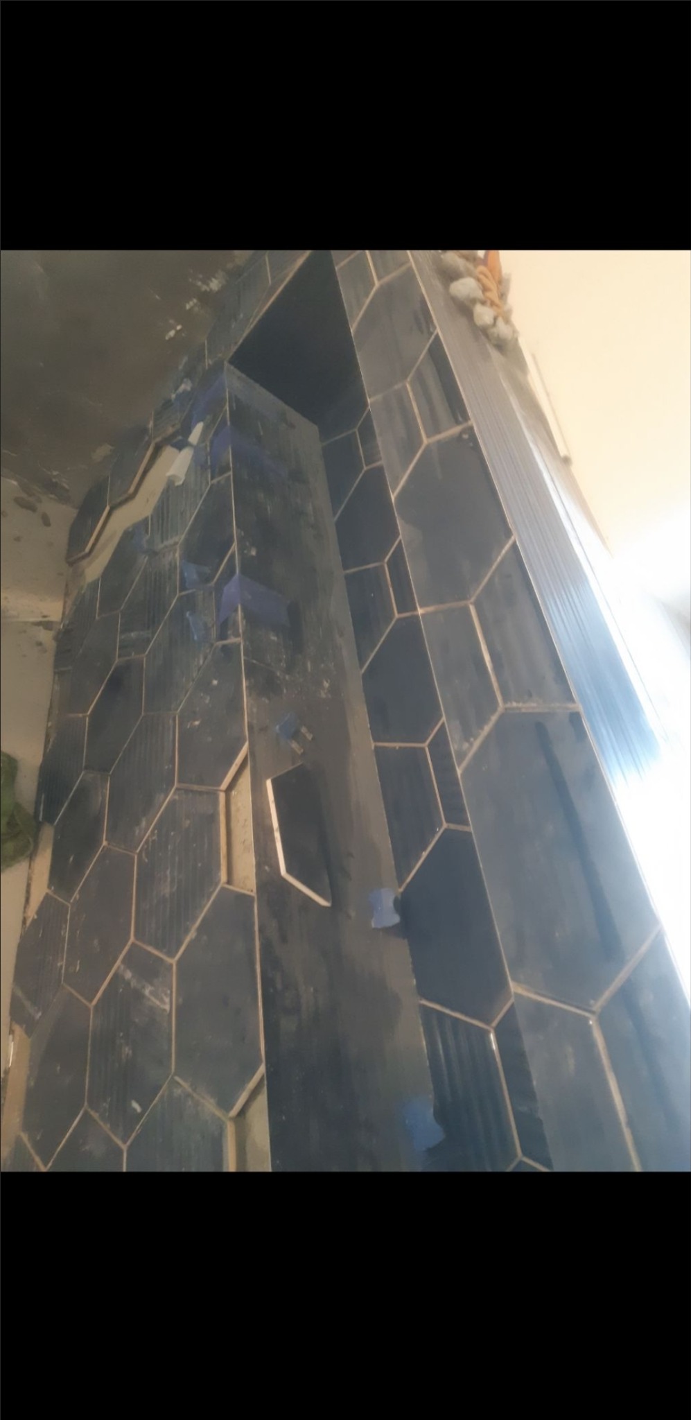 Układanie heksagonalnych, ciemnych płytek na ścianie, widoczne niebieskie kliny dystansowe, fragment surowego tynku i pianki montażowej.