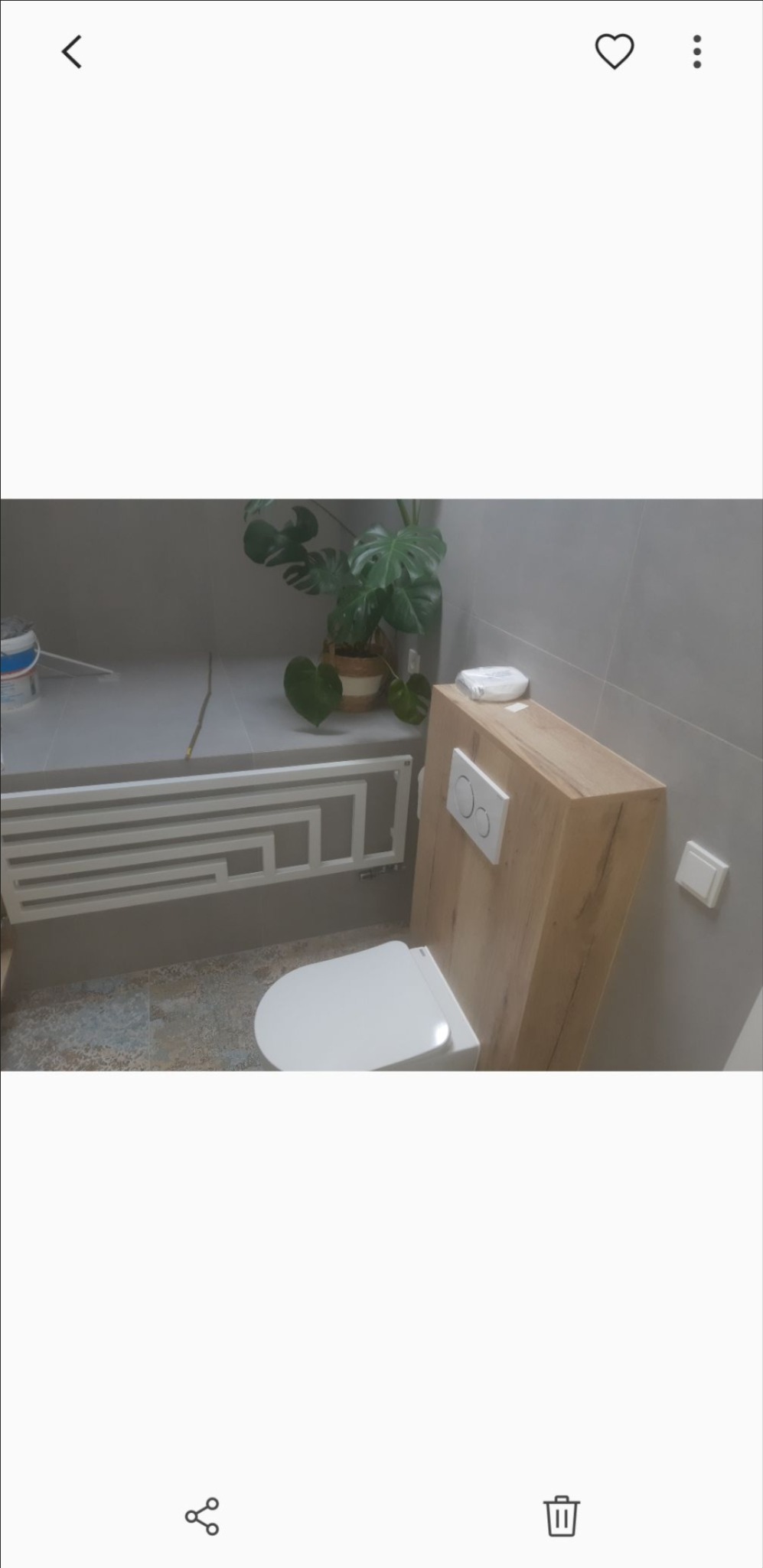 Nowoczesna łazienka z podwieszaną toaletą obudowaną drewnopodobną płytą, dekoracyjnym grzejnikiem i rośliną Monstera w doniczce.