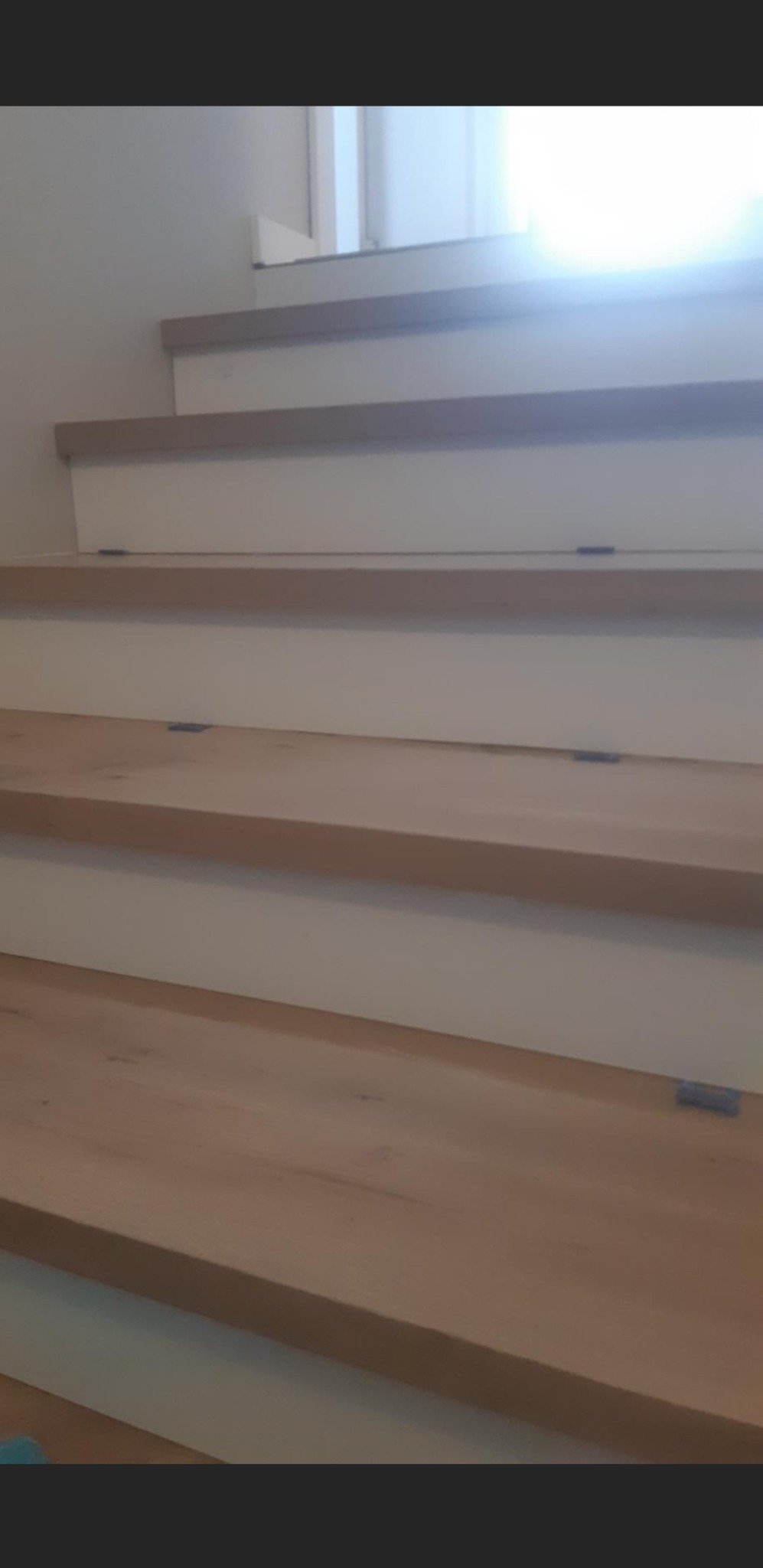 Drewniane schody z białymi podstopnicami, widoczne metalowe mocowania stopni, jasne światło z okna w tle.