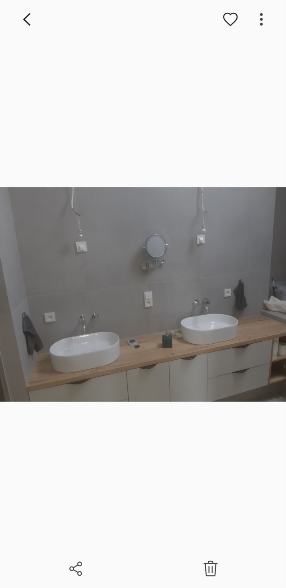 Dwie owalne umywalki nablatowe na drewnianym blacie z szufladami w łazience wykończonej szarymi płytkami, widoczne niedokończone podłączenia elektryczne i małe okrągłe lusterko.