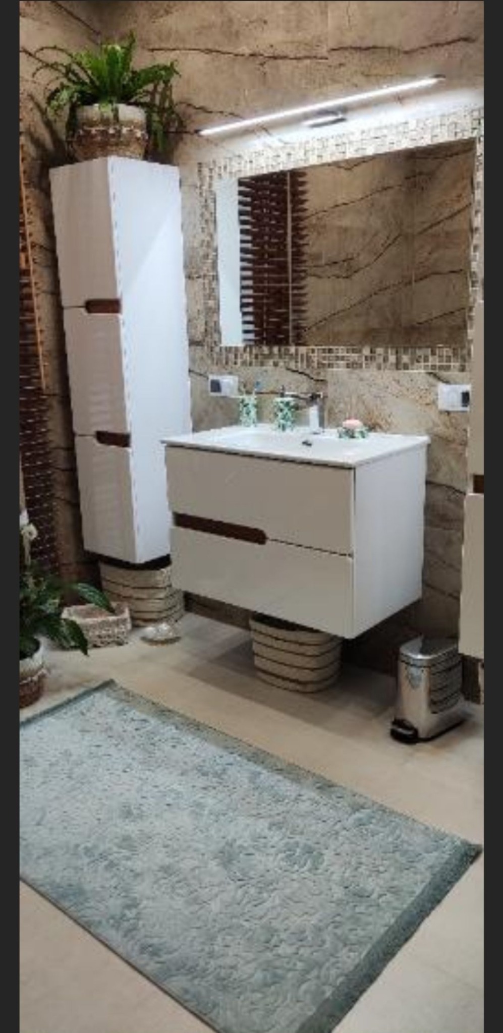 Nowoczesna łazienka z białymi szafkami pod umywalką i wysoką szafką boczną, obie z ciemnymi uchwytami, ustawione na plecionych koszach, ściany wykończone płytkami imitującymi kamień, dywanik...