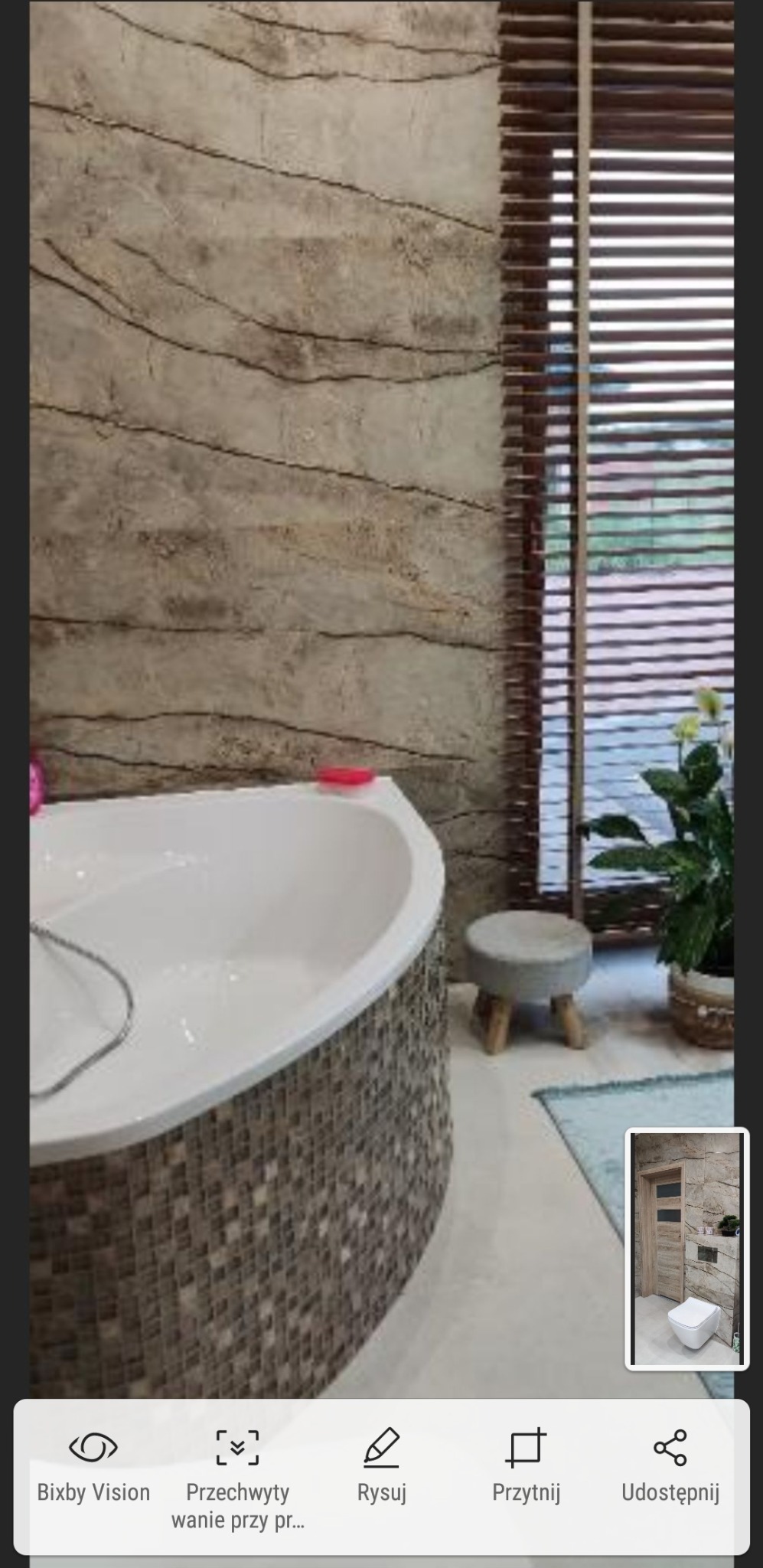 Biała wanna z mozaikową obudową na tle ściany imitującej kamień i drewnianych żaluzji okiennych, uzupełniona o stołek i roślinę doniczkową.