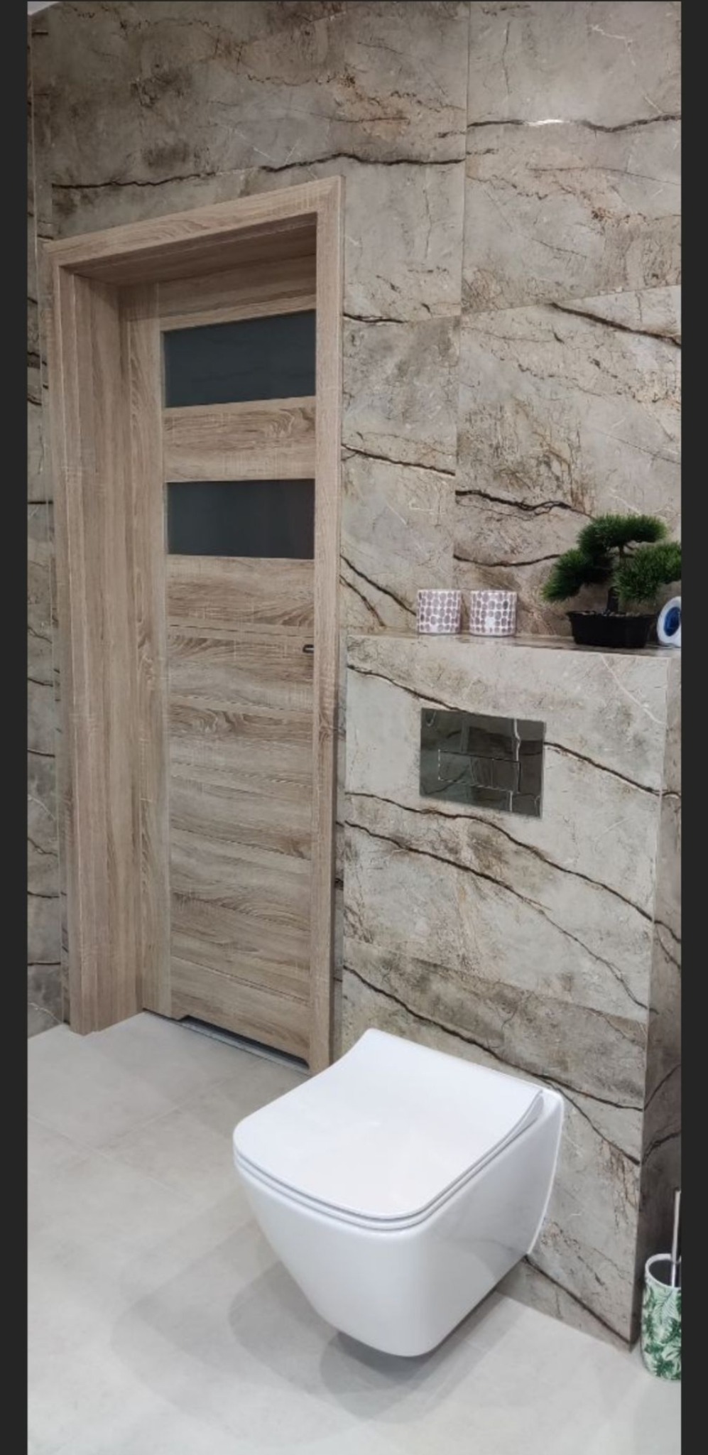 Nowoczesna toaleta podwieszana w łazience wykończonej płytkami imitującymi marmur, z drewnianymi drzwiami z przeszkleniami i elementami dekoracyjnymi na półce.