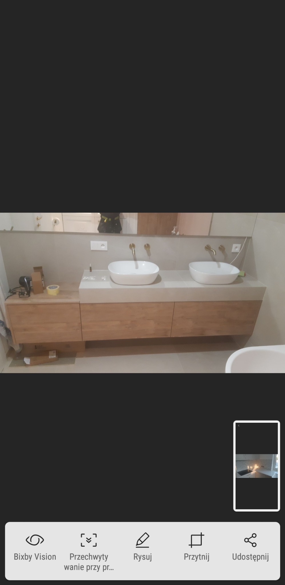 Nowoczesna łazienka z dwoma białymi umywalkami nablatowymi i złotymi bateriami, drewniana szafka pod umywalką, jasne płytki na ścianie, widoczne akcesoria i kosmetyki.