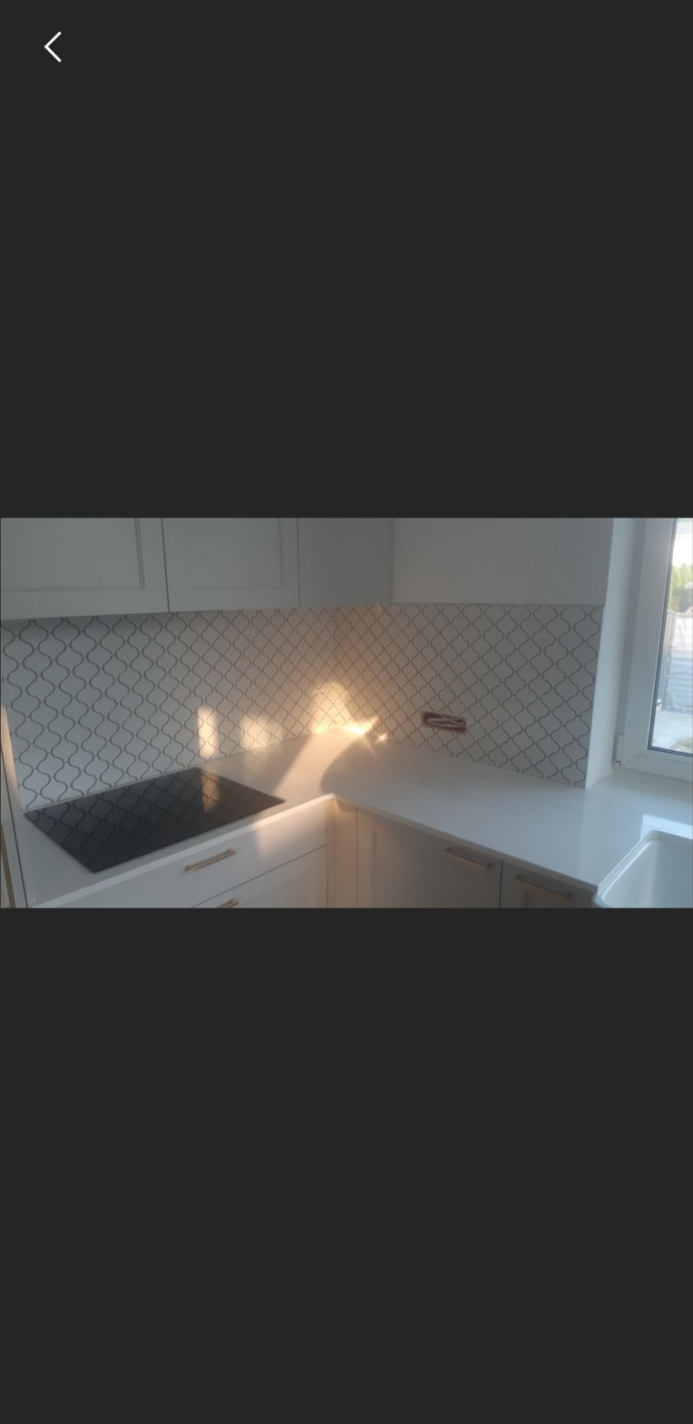 Jasna kuchnia z białymi szafkami, złotymi uchwytami i dekoracyjną mozaiką na ścianie nad blatem, oświetlona promieniami słonecznymi wpadającymi przez okno.