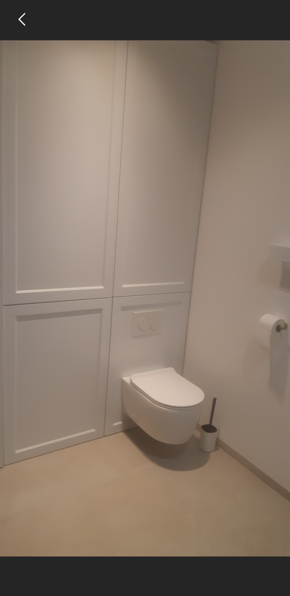 Nowoczesna, podwieszana toaleta w białej obudowie, z ukrytym spłukiwaniem i szczotką do WC, w minimalistycznej łazience z beżową podłogą.