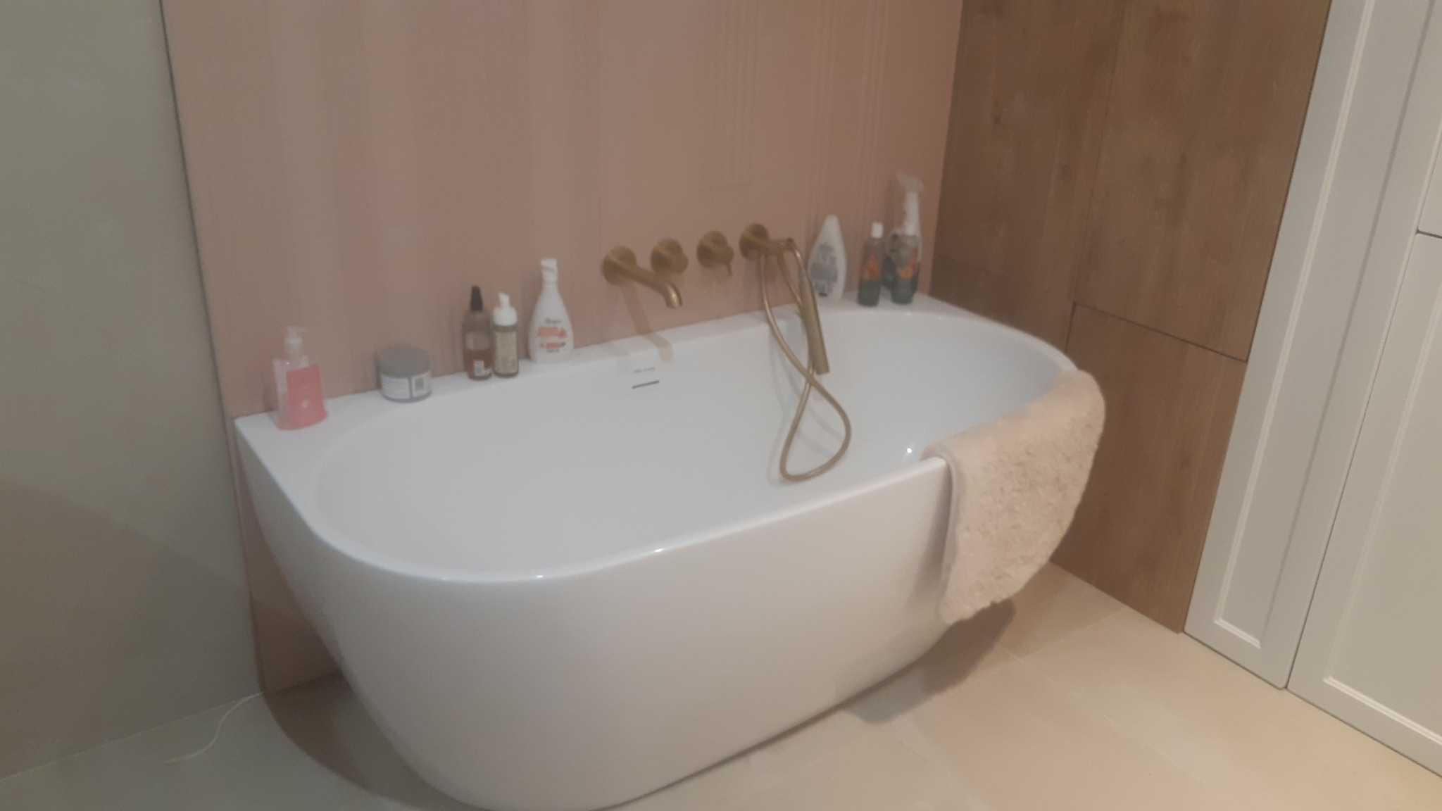 Wolnostojąca, biała wanna z armaturą w kolorze szczotkowanego złota, z kosmetykami na krawędzi i beżowym ręcznikiem, na tle ściany w odcieniach beżu i różu oraz drewnopodobnej zabudowy.