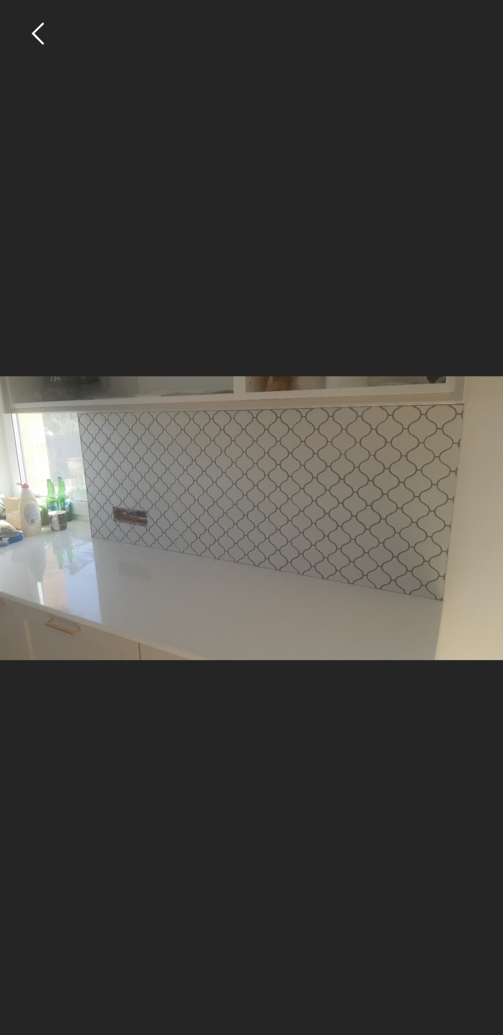 Kuchnia z błyszczącym, białym blatem i płytkami ściennymi o wzorze marokańskiej koniczyny w odcieniach szarości, widoczne butelki na blacie i białe szafki.