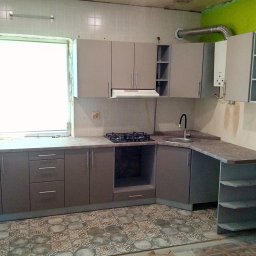 Aleksandr Vasilenko - Szara zabudowa kuchenna z gazową płytą grzewczą i zlewozmywakiem, widok na okno i fragment ściany z zielonym akcentem, wzorzysta podłoga.