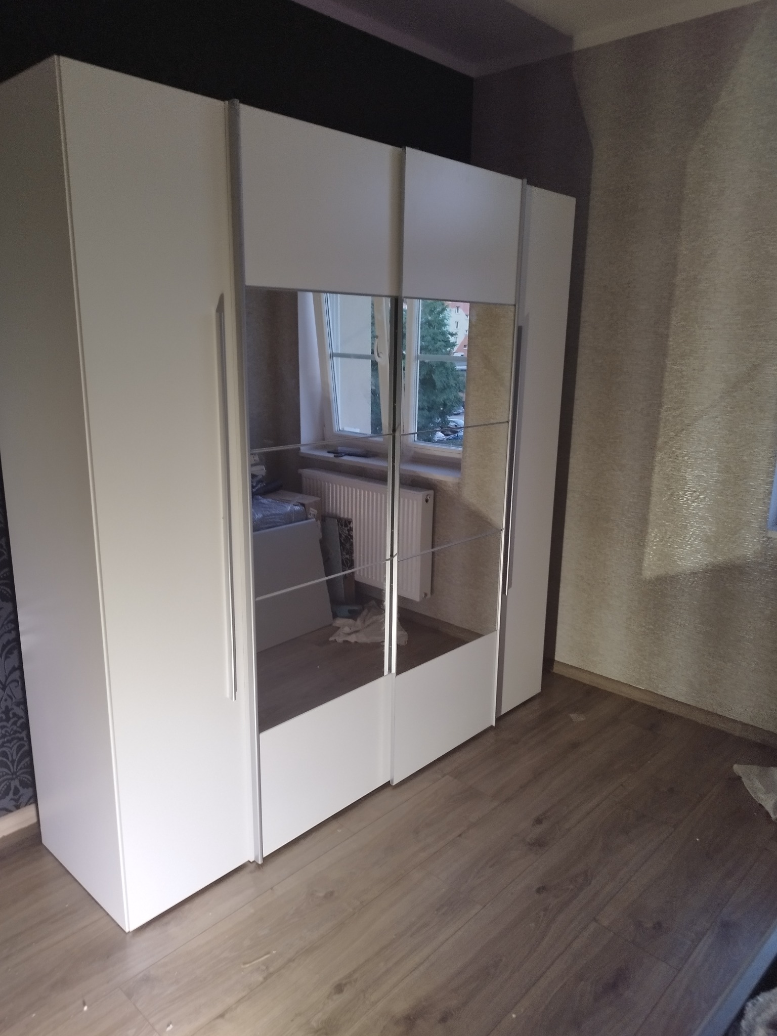 Biała szafa przesuwna z lustrami w pokoju z parkietem, widoczne odbicie okna i grzejnika.