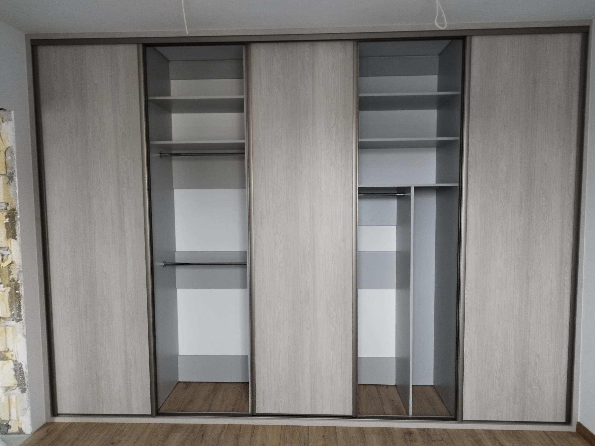 Szafa przesuwna z jasnego drewna z widocznymi półkami i drążkami na ubrania, częściowo otwarta, na podłodze panele.