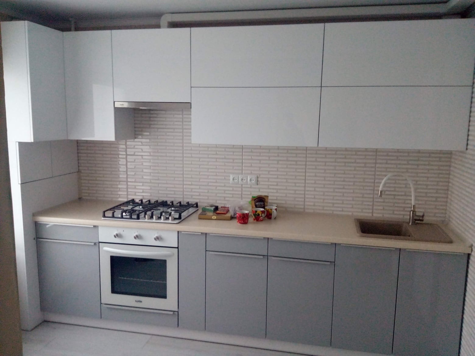 Szara zabudowa kuchenna z białymi szafkami górnymi i beżowym blatem, widoczna płyta gazowa z palnikami, zlewozmywak z baterią i płytki imitujące cegłę na ścianie.