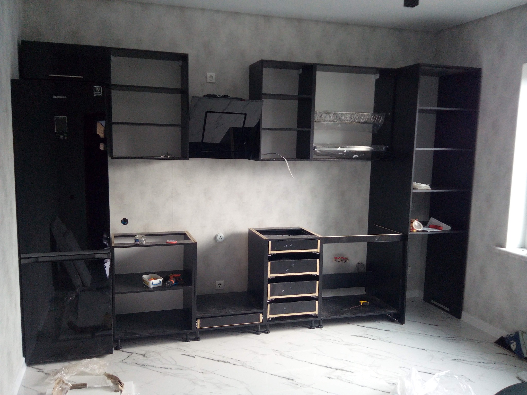 Częściowo zmontowana, czarna zabudowa kuchenna z lodówką Samsung, widoczne narzędzia i akcesoria montażowe na marmurowej podłodze.