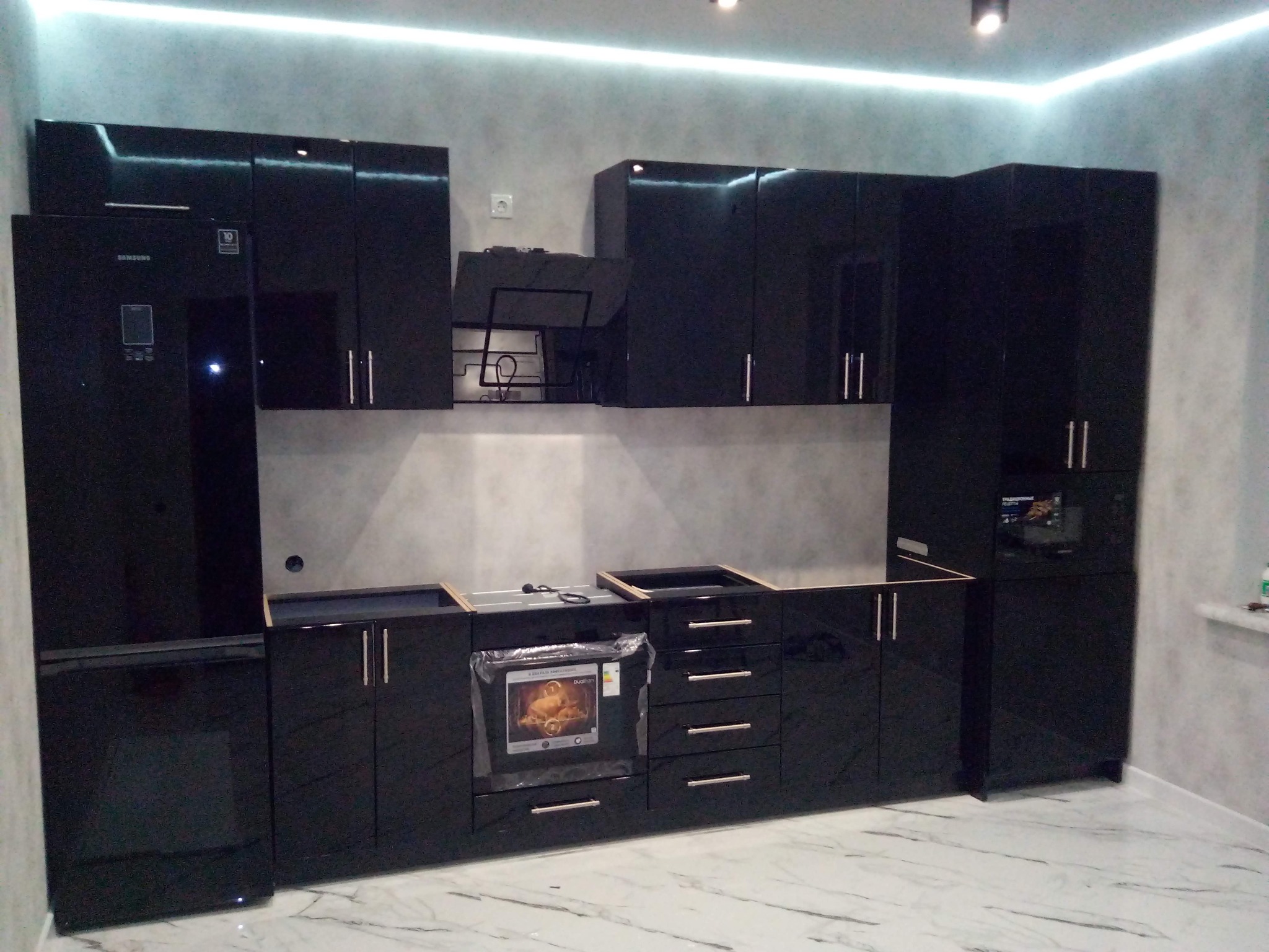 Nowoczesna, czarna, lakierowana zabudowa kuchenna z lodówką Samsung, płyta grzewcza i piekarnik w folii ochronnej, na podłodze marmurkowy wzór, oświetlenie LED podszafkowe.