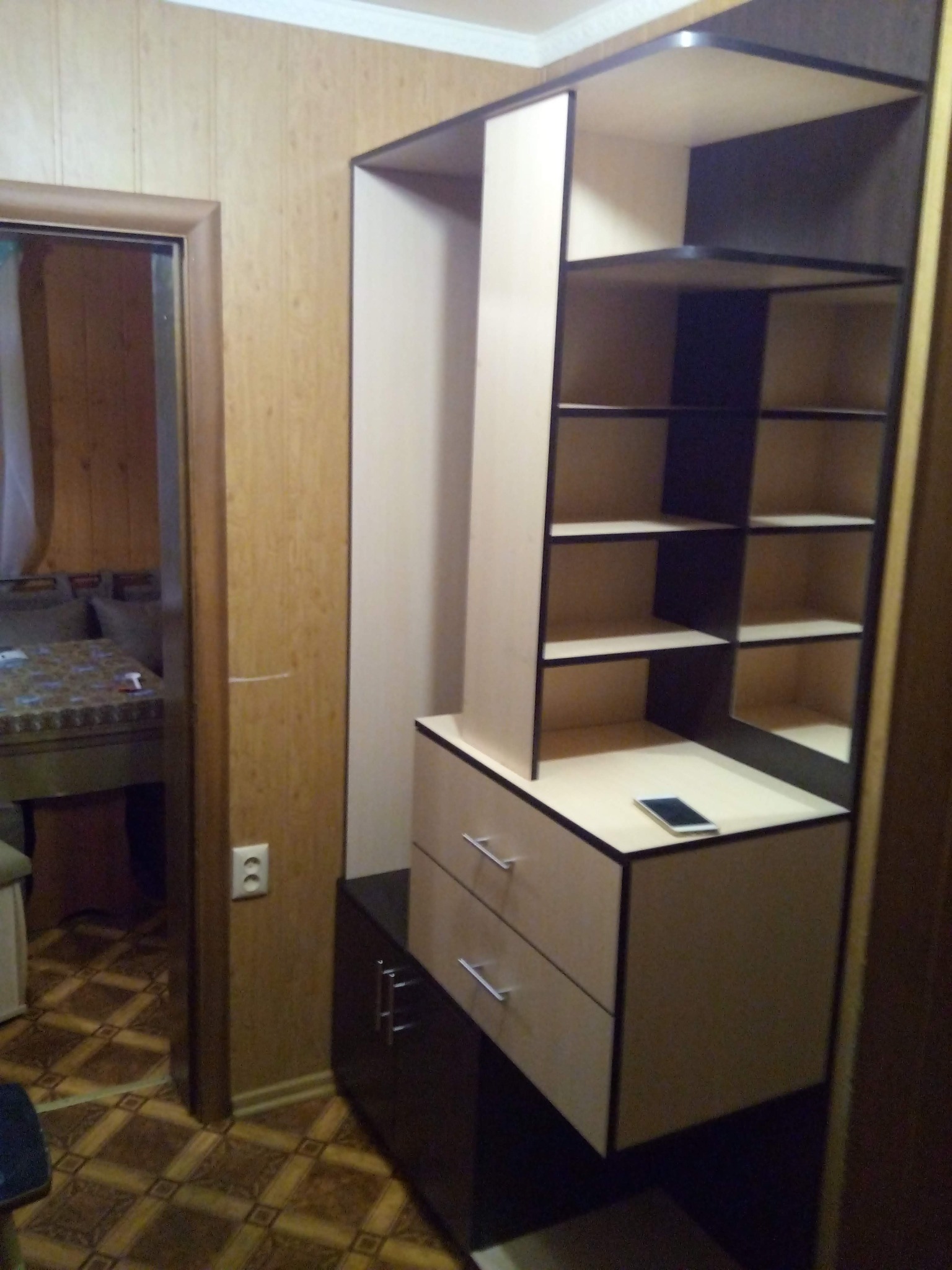 Wysoka szafa z szufladami i półkami w brązowo-beżowej kolorystyce, z telefonem na blacie, ustawiona w rogu pokoju z boazerią i wzorzystą podłogą.