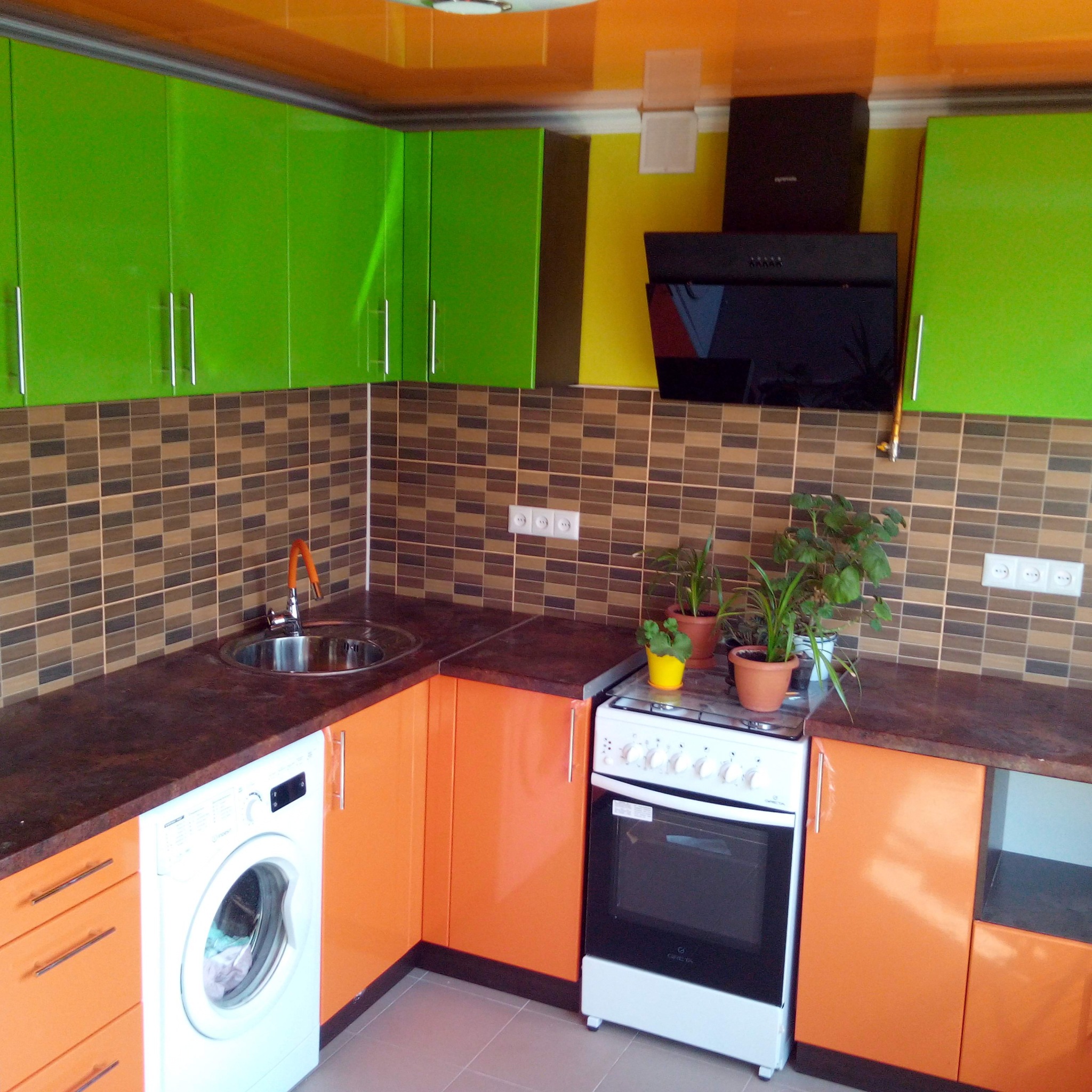 Narożna kuchnia z zielonymi szafkami wiszącymi i pomarańczowymi szafkami dolnymi, z pralką w zabudowie i kuchenką, wykończona brązowymi płytkami mozaikowymi.