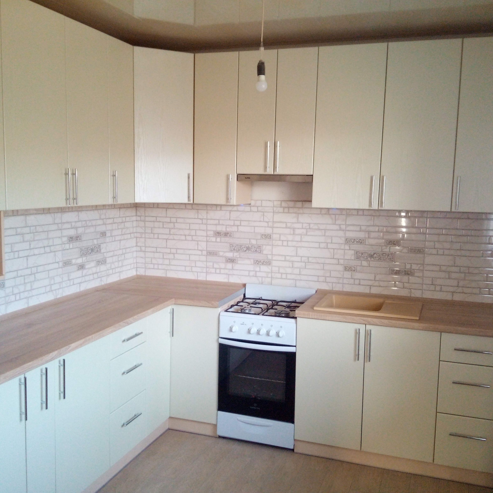 Jasna kuchnia z beżowymi szafkami, drewnianym blatem i płytkami imitującymi cegłę z dekoracyjnymi wstawkami, z białą kuchenką gazową i wiszącą żarówką bez klosza.