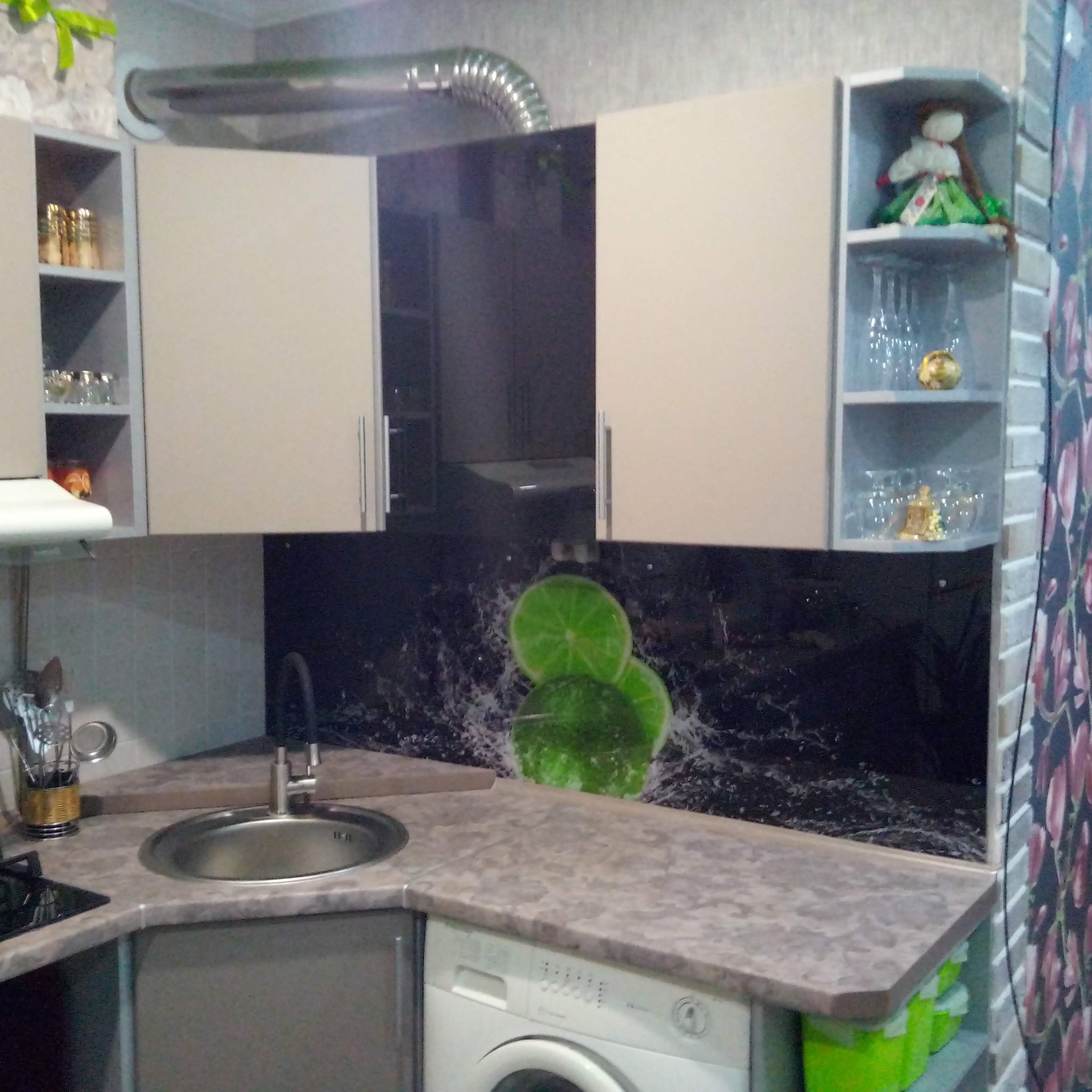 Komplet szafek kuchennych w rogu pomieszczenia z blatem imitującym marmur, zlewozmywakiem i panelem ściennym z motywem limonki oraz widoczną pralką.