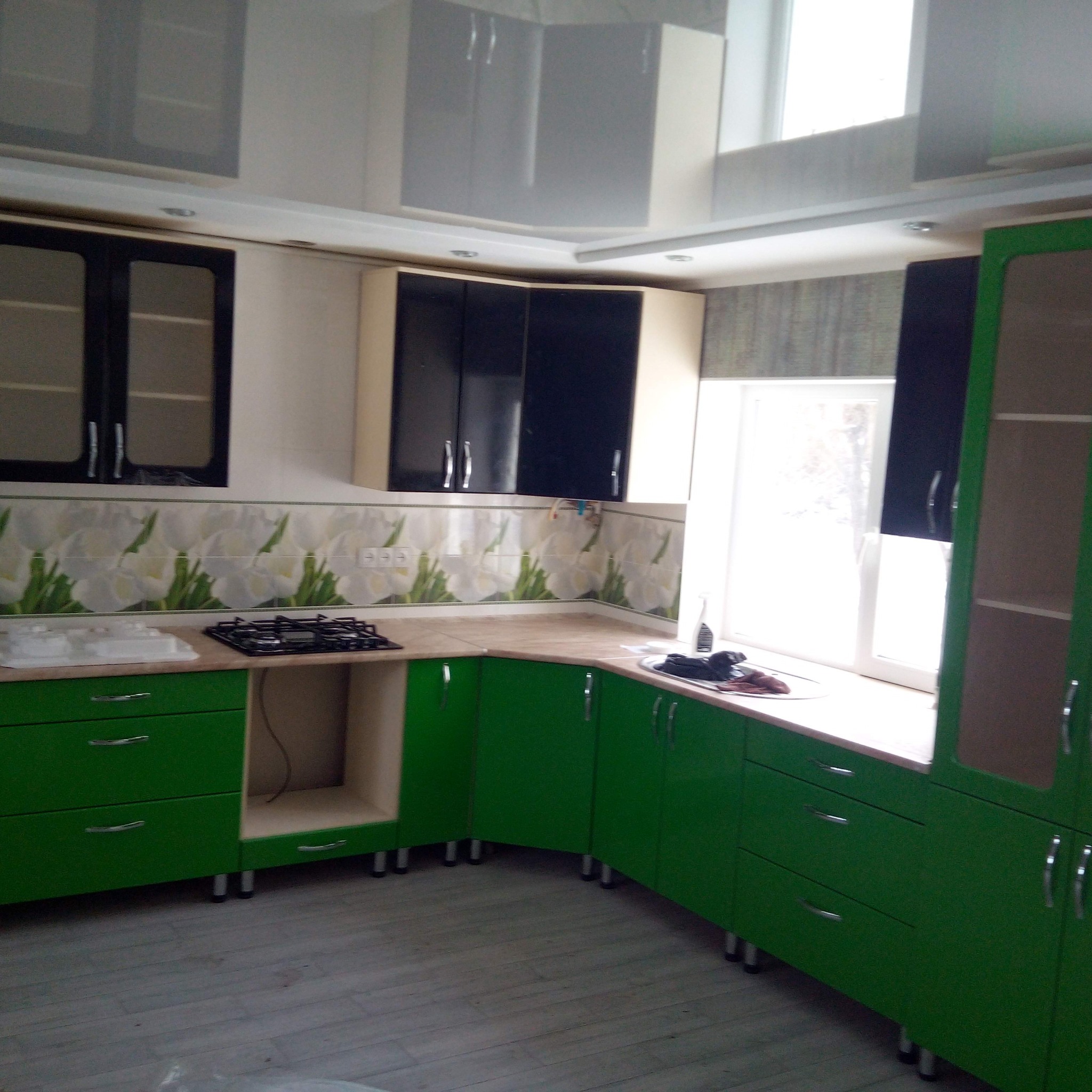 Nowoczesna kuchnia z zielonymi szafkami, czarnymi górnymi szafkami i jasnym blatem, wykończona płytkami z motywem kwiatowym, oświetlona światłem dziennym wpadającym przez okno.