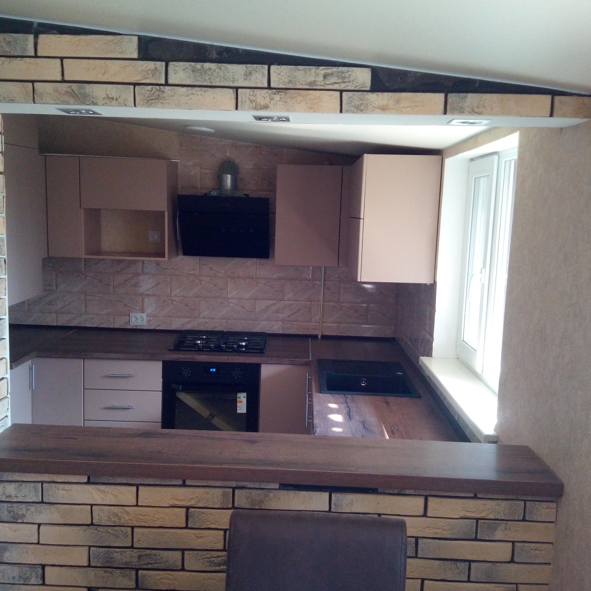 Wykończona kuchnia z beżowymi szafkami, czarnym okapem i blatem z imitacją drewna, widoczna przez otwór w ścianie wyłożonej cegłą.