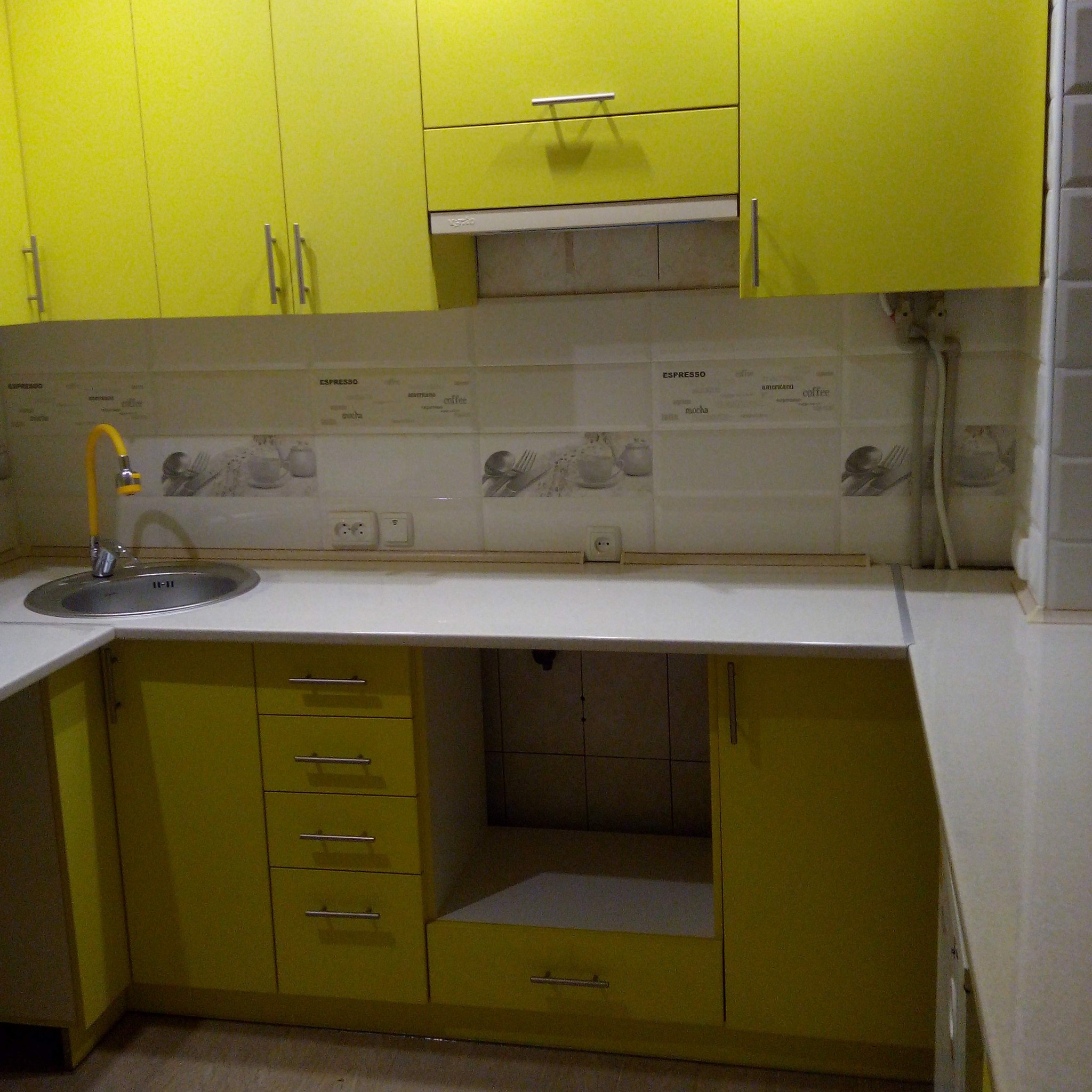 Żółte szafki kuchenne z chromowanymi uchwytami, biały blat, żółty kran i płytki z motywem kawy.