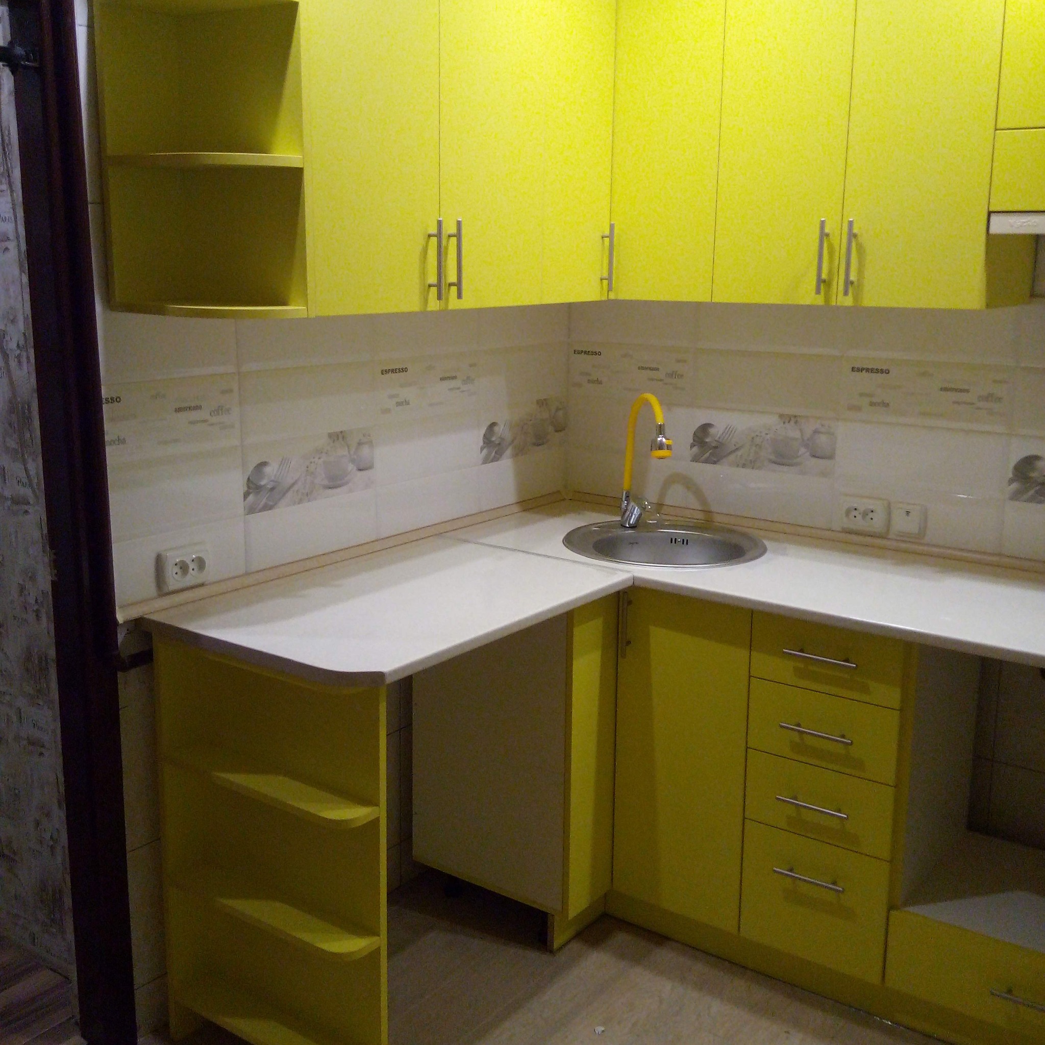 Żółta zabudowa kuchenna z białym blatem, chromowanym zlewem i baterią. Na ścianie płytki z motywem kawy. Widoczne gniazdka elektryczne.