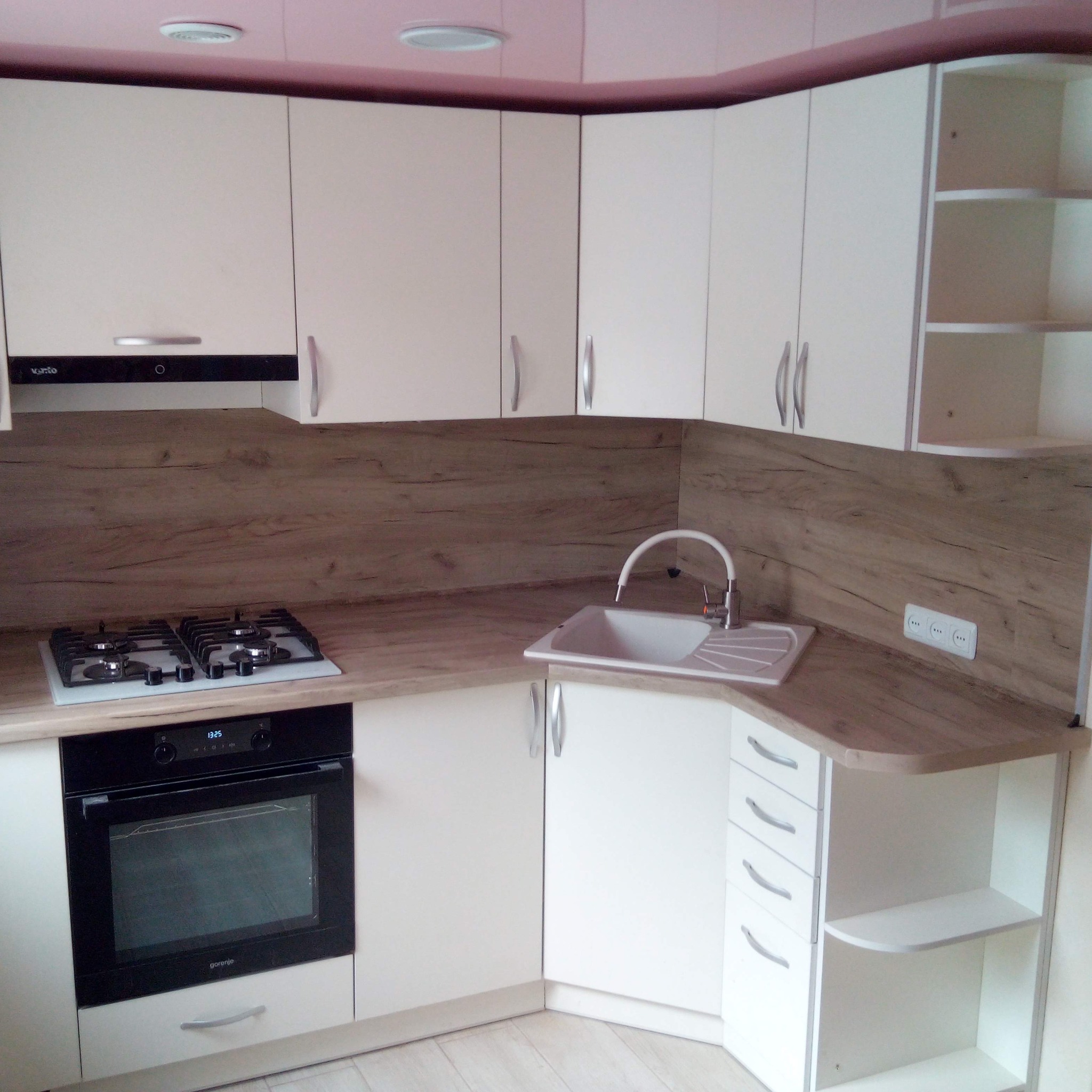 Nowoczesna kuchnia narożna z białymi szafkami, drewnopodobnym blatem i sprzętem AGD: piekarnikiem Gorenje i płytą gazową, z widocznym oświetleniem sufitowym i gniazdkiem elektrycznym.