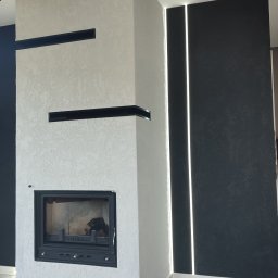 Home-bud - Nowoczesny kominek obudowany jasnym tynkiem strukturalnym z czarnymi wnękami, sąsiadujący z ciemną ścianą z pionowymi paskami ledowymi, na marmurowej podłodze.