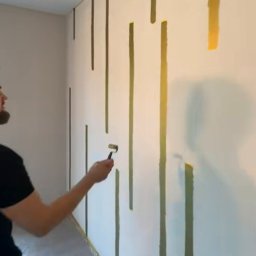 Home-bud - Malowanie ściany wałkiem na złoto w pionowe pasy, widoczny fragment pomieszczenia zabezpieczonego folią malarską.