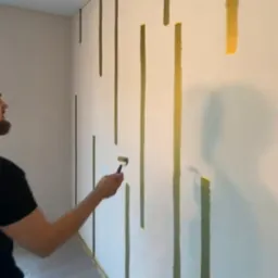 Malowanie ściany wałkiem na złoto w pionowe pasy, widoczny fragment pomieszczenia zabezpieczonego folią malarską.