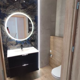 Home-bud - Nowoczesna łazienka w Tychach z okrągłym lustrem podświetlanym LED, czarną szafką pod umywalką i drewnopodobnymi płytkami na podłodze.