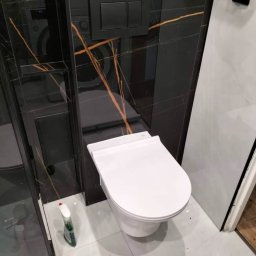 Home-bud - Biała, podwieszana toaleta z czarnym panelem spłukującym na ścianie wyłożonej czarnymi płytkami z pomarańczowymi żyłami, widoczna kabina prysznicowa i butelka z płynem do czyszczenia na białej...