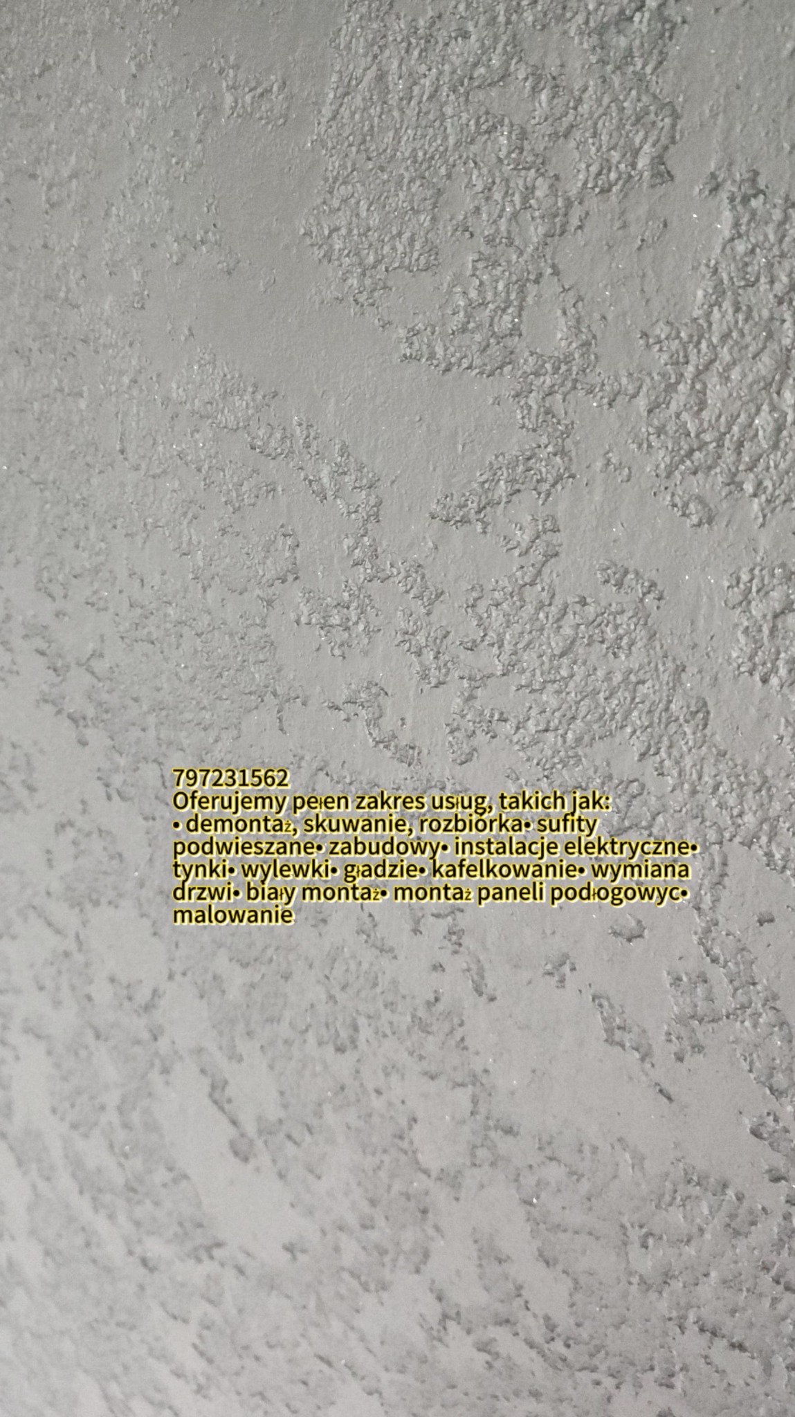 Szorstka, biała ściana z widocznym tekstem reklamowym naklejonym na dolnej części, oferującym szeroki zakres usług budowlanych, w tym demontaż, skuwanie, sufity podwieszane, instalacje elektryczne,...