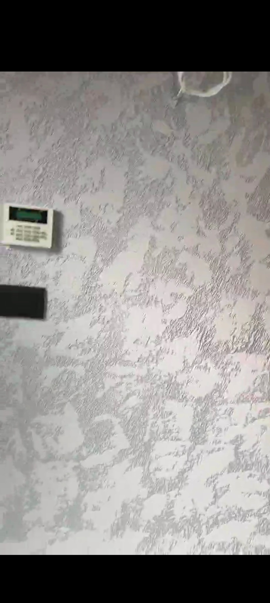 Ściana pokryta dekoracyjnym tynkiem w odcieniach bieli i srebra, z zamontowanym panelem alarmowym i wystającym kablem.