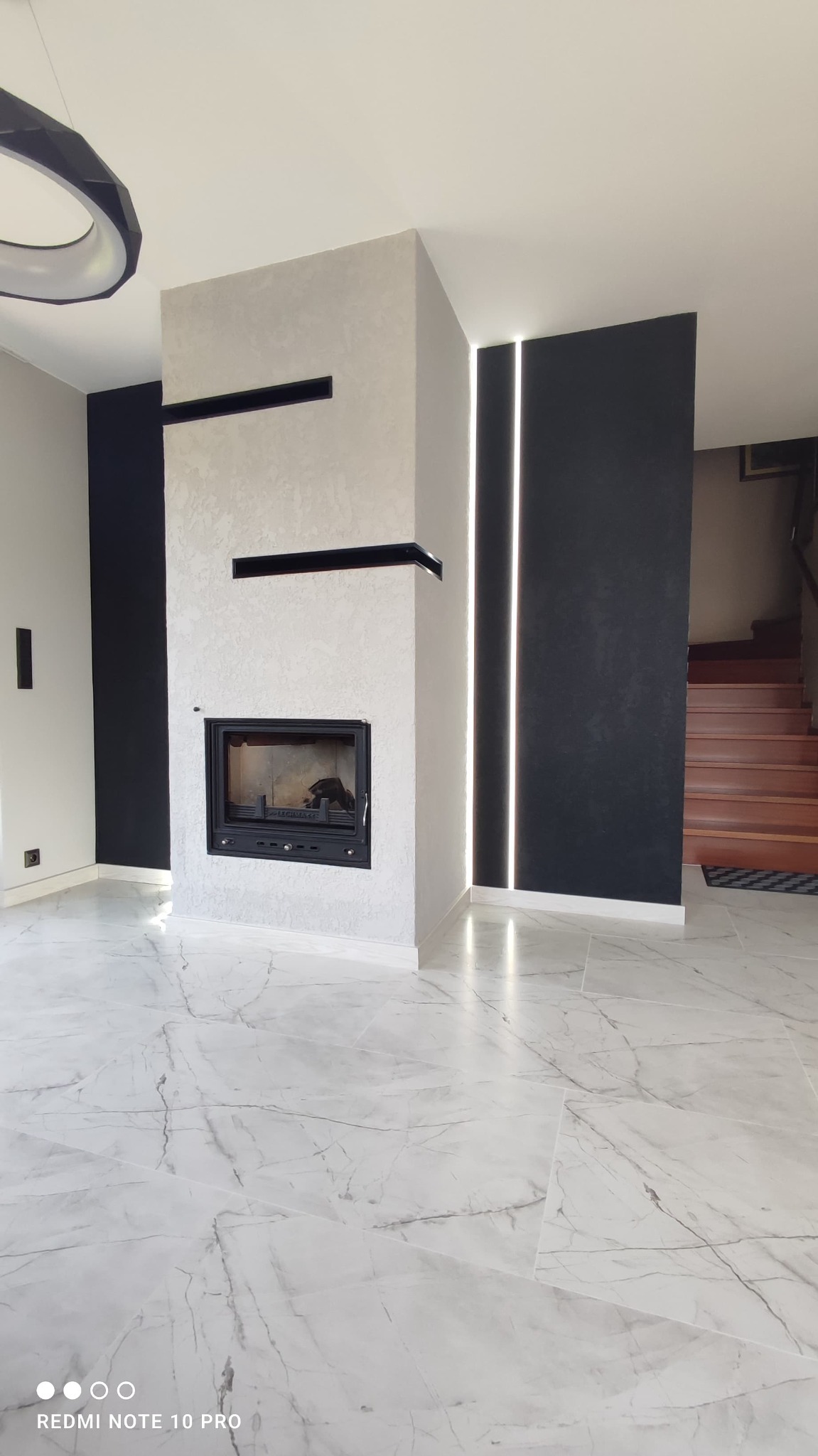 Nowoczesny salon z kominkiem obudowanym w jasny tynk strukturalny, czarne wnęki dekoracyjne, podświetlane pionowe pasy LED na czarnej ścianie, podłoga z płytek imitujących biały marmur z szarymi...