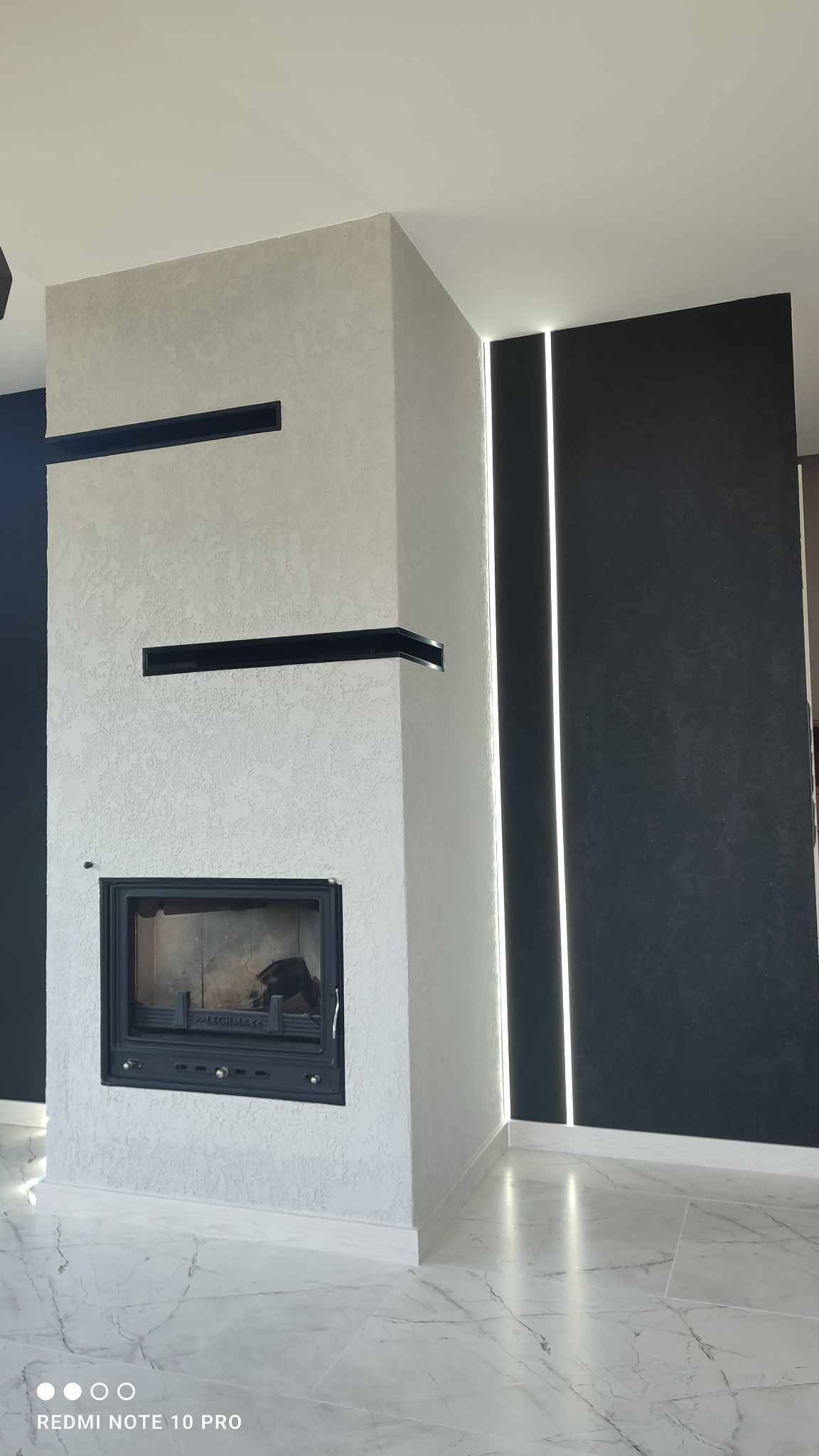 Nowoczesny kominek obudowany jasnym tynkiem strukturalnym z czarnymi wnękami, sąsiadujący z ciemną ścianą z pionowymi paskami ledowymi, na marmurowej podłodze.