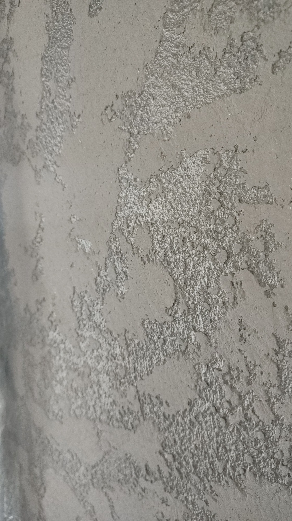 Szara ściana z dekoracyjną, nierównomierną fakturą, przypominającą efekt stiuku weneckiego, z wyraźnymi wklęsłościami i wypukłościami.