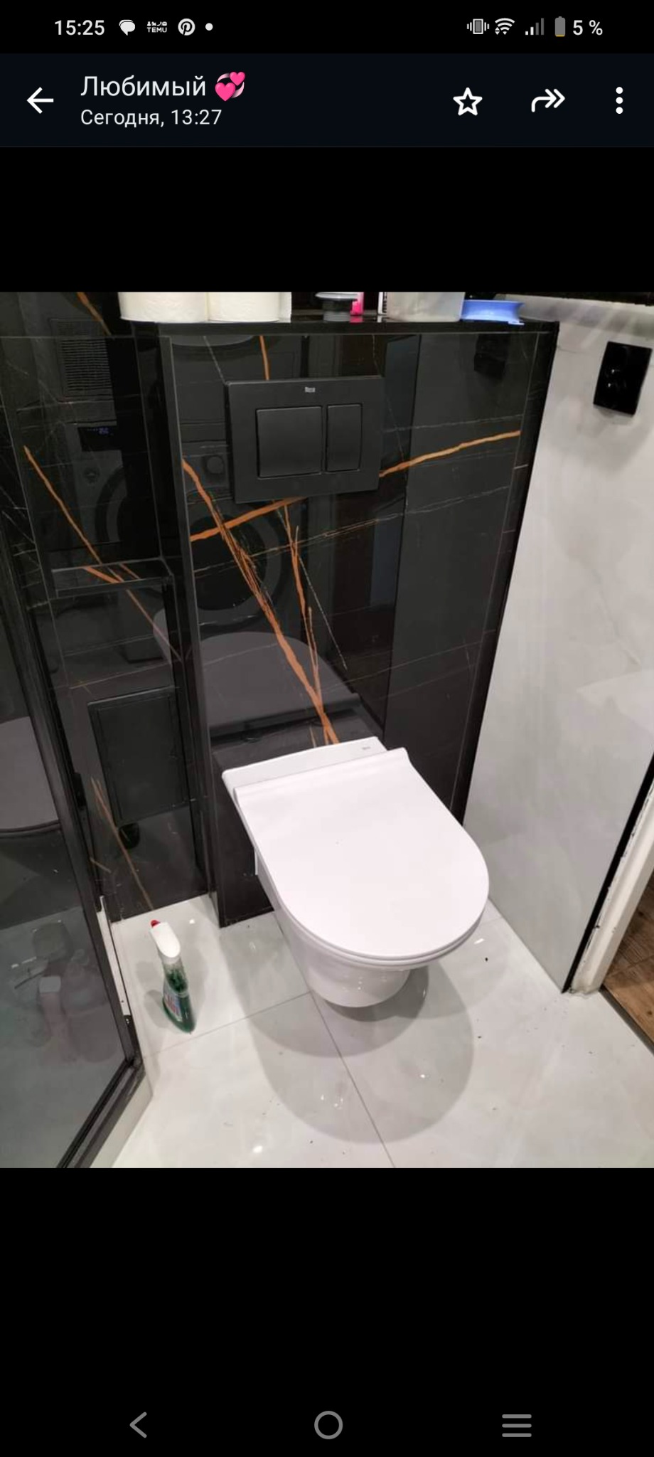 Biała, podwieszana toaleta z czarnym panelem spłukującym na ścianie wyłożonej czarnymi płytkami z pomarańczowymi żyłami, widoczna kabina prysznicowa i butelka z płynem do czyszczenia na białej...