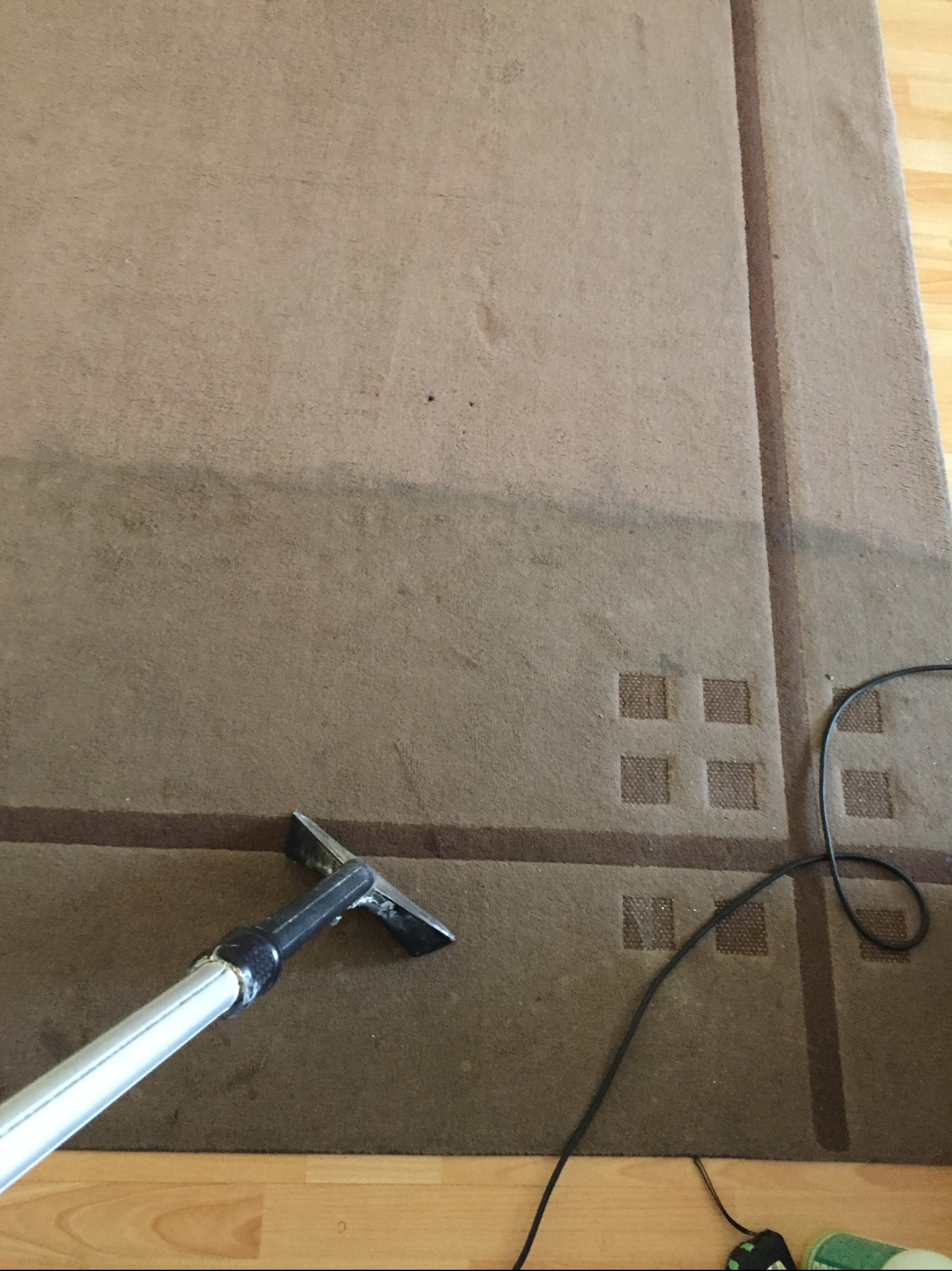 Fragment brązowego dywanu z geometrycznym wzorem, w trakcie czyszczenia odkurzaczem piorącym; widoczna różnica w kolorze pomiędzy wyczyszczoną a nie wyczyszczoną powierzchnią.